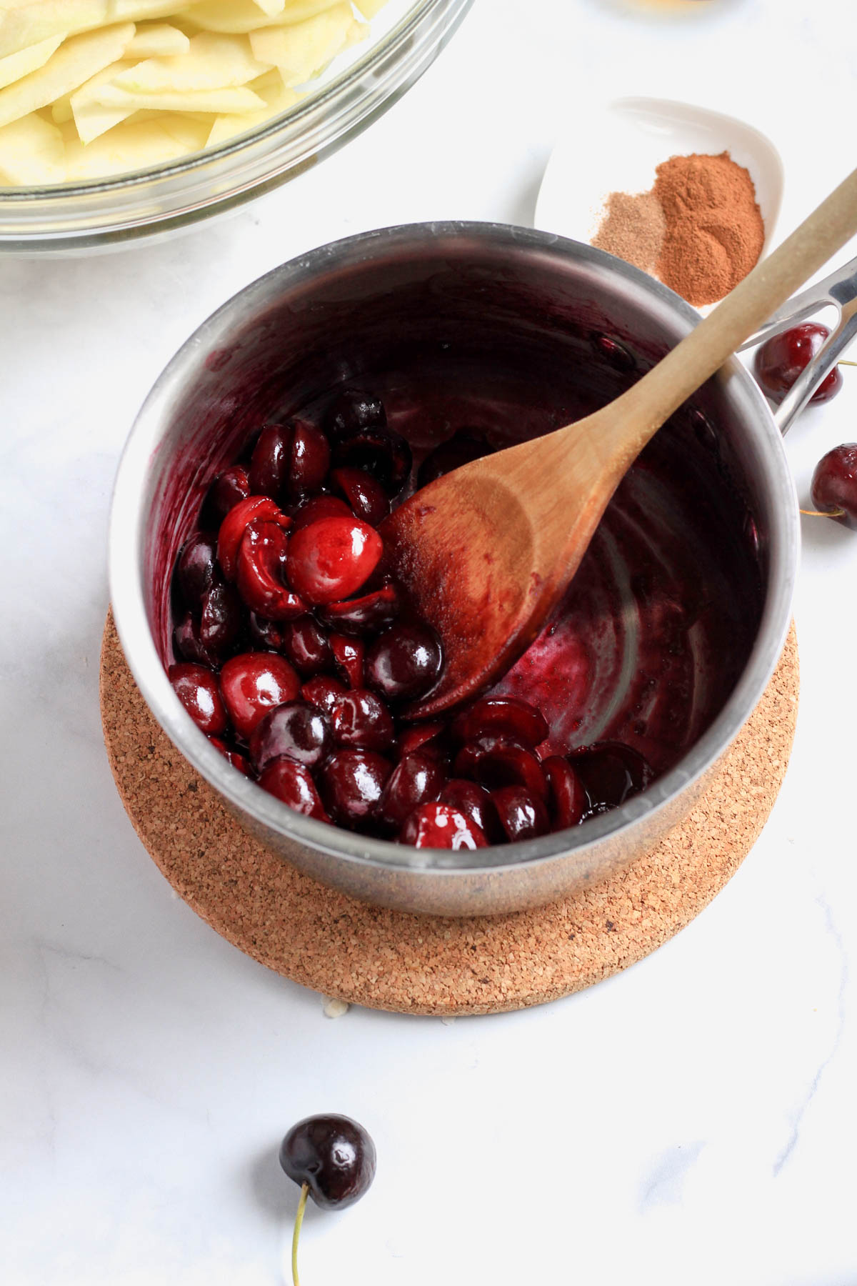 A silver sauce pan with a wooden spoon and cooked cherries with juices running on a brown cork trivet.