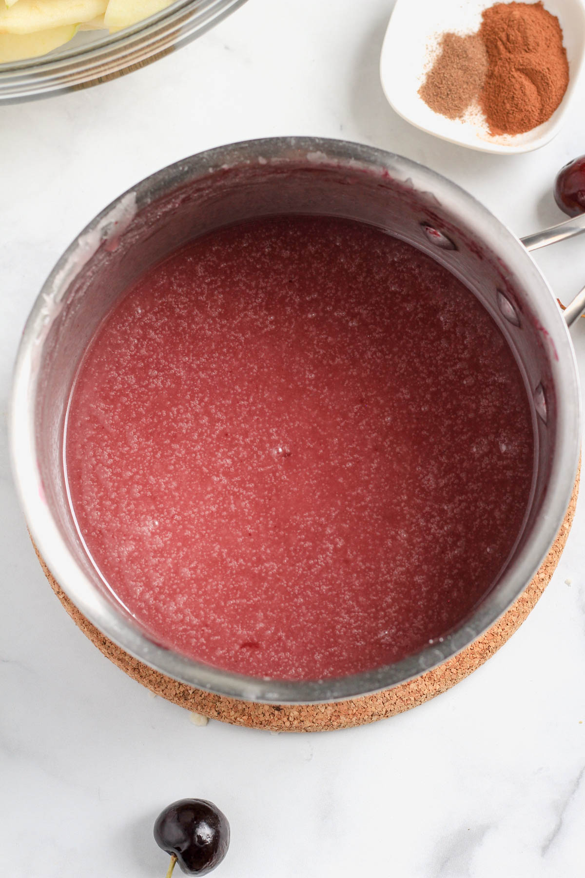 A silver sauce pan with the cherry juice filling on a brown cork trivet.