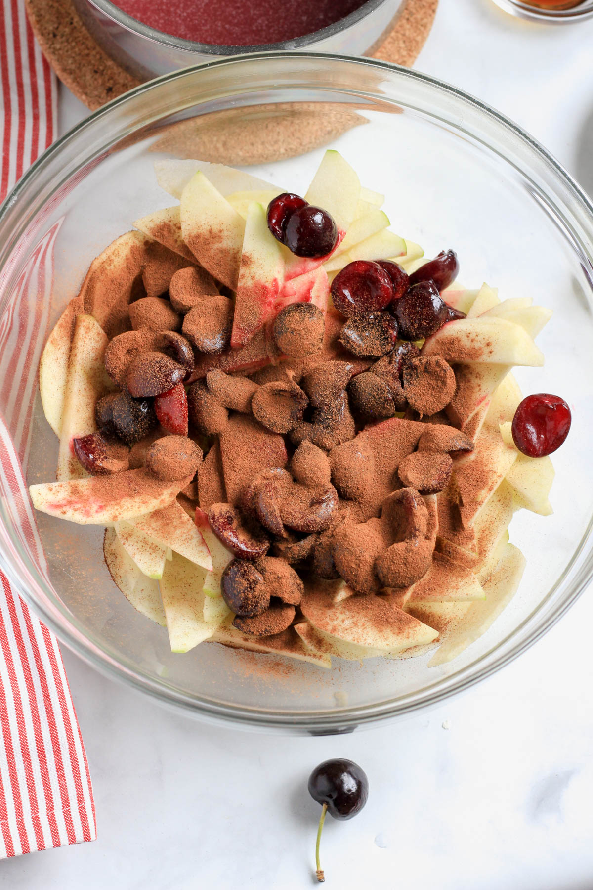 A glass bowl with apple slices and cherries topped with cinnamon and all spice before mixing to coat.