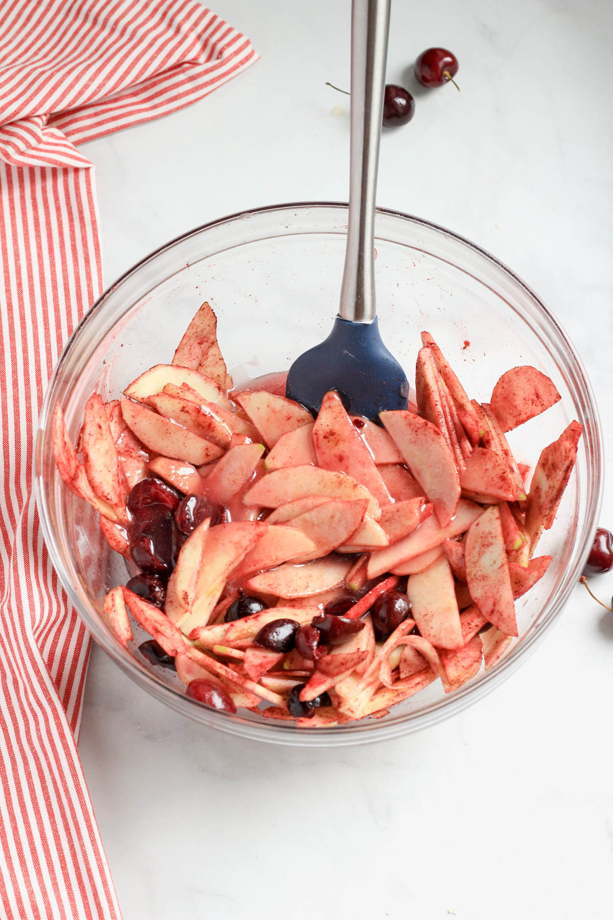 A glass bowl with a blue and silver spatula stirring in the cherry apple pie filling.