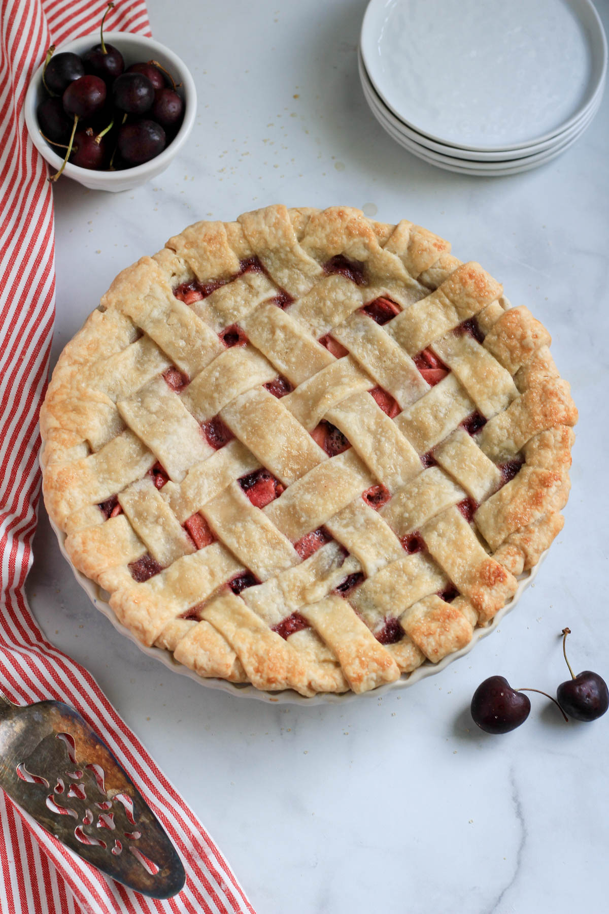 Vegan cherry apple pie after baking on a white counter with a red and white striped dish towel to the left.