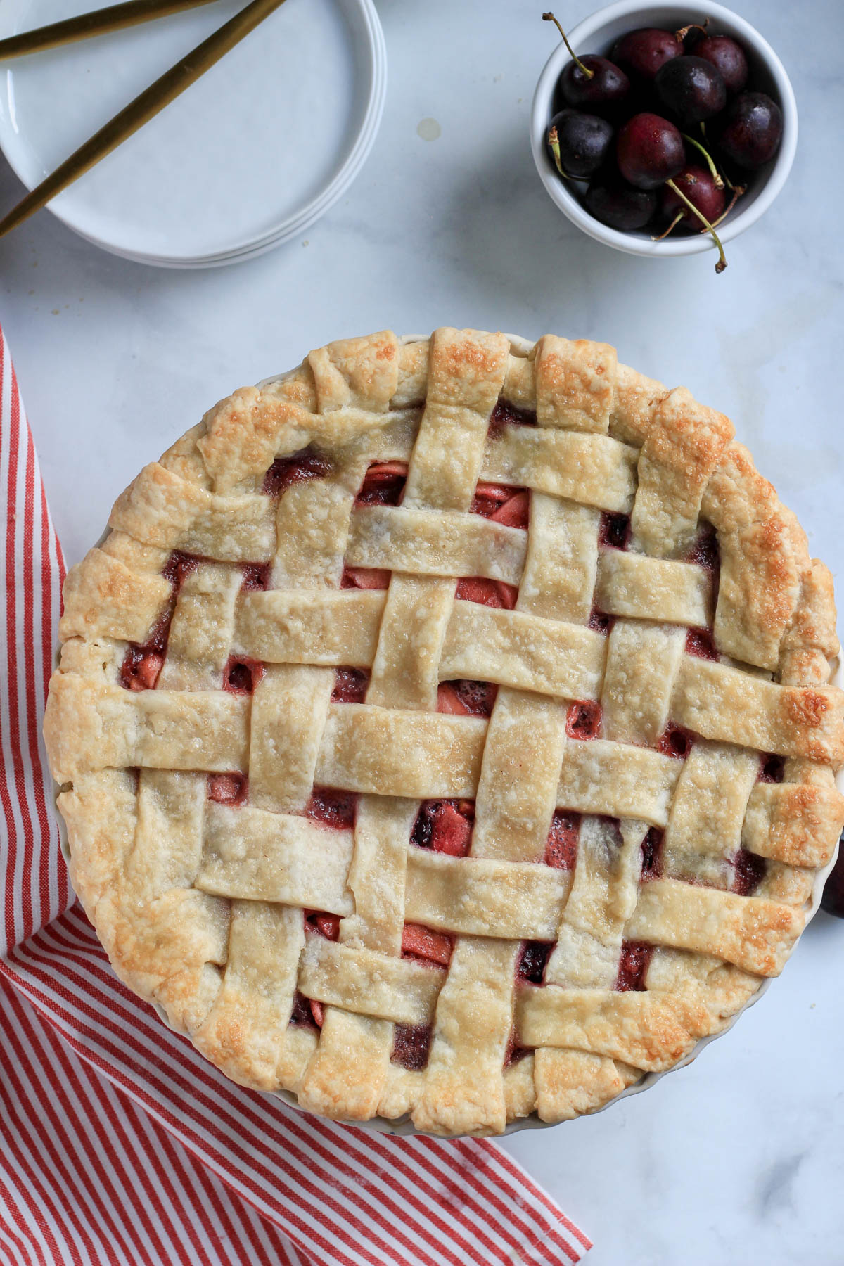 Vegan cherry apple pie in a pie pan with a bowl of cherries in the top right and a stack of white plates in the top left.