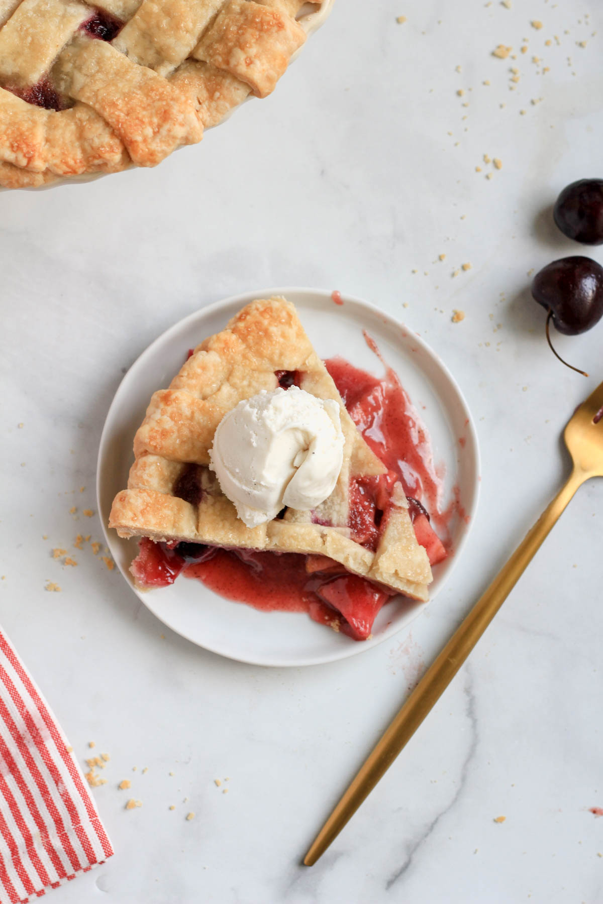 A slice of cherry apple pie topped with vegan ice cream with a gold fork to the right of the plate.