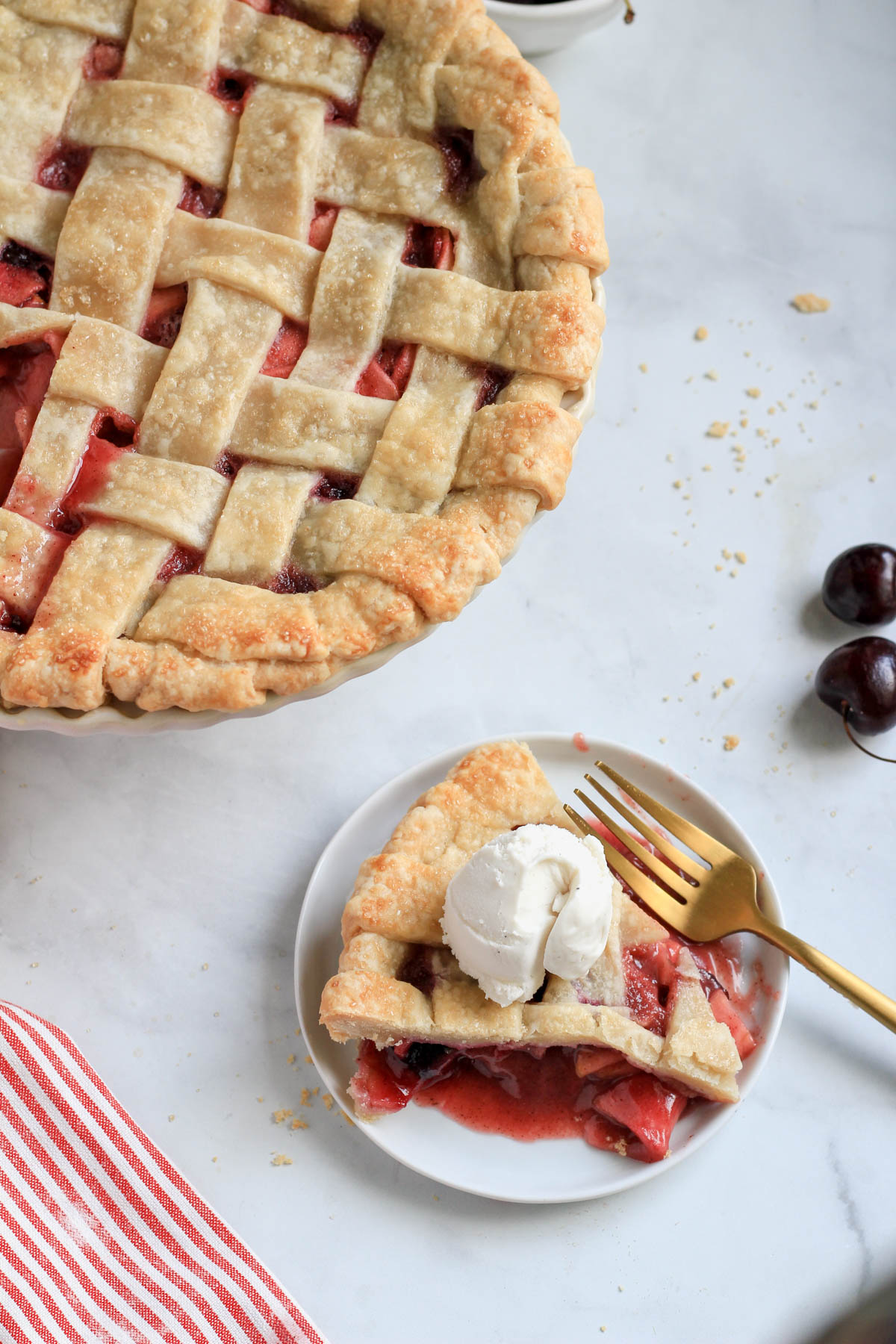 A small white plate of cherry apple pie with a gold fork topped with ice cream and a full cherry apple pie in the top left corner.
