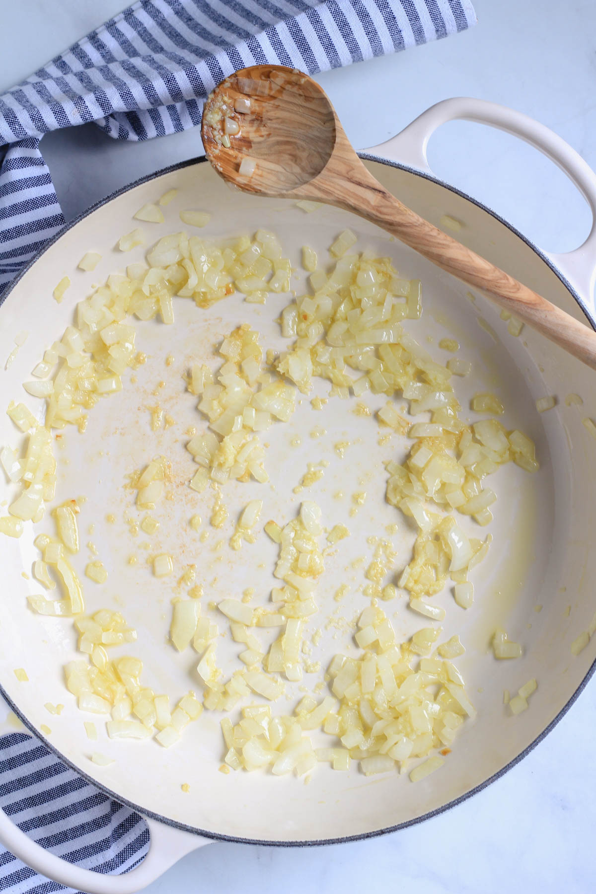 A white skillet with onions and garlic and a wooden spoon.