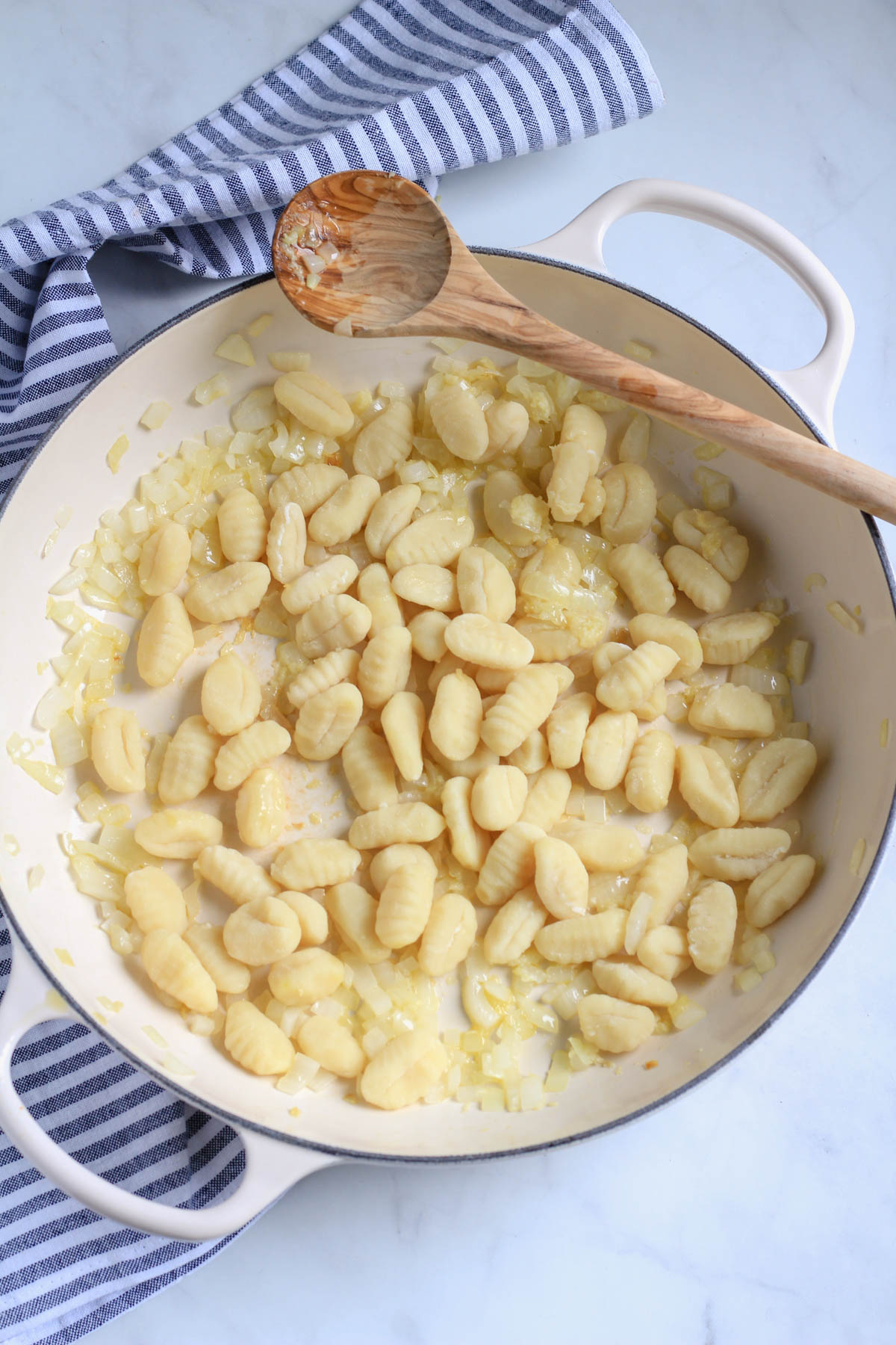 A white skillet with gnocchi, onions, and garlic, with a wooden spoon.