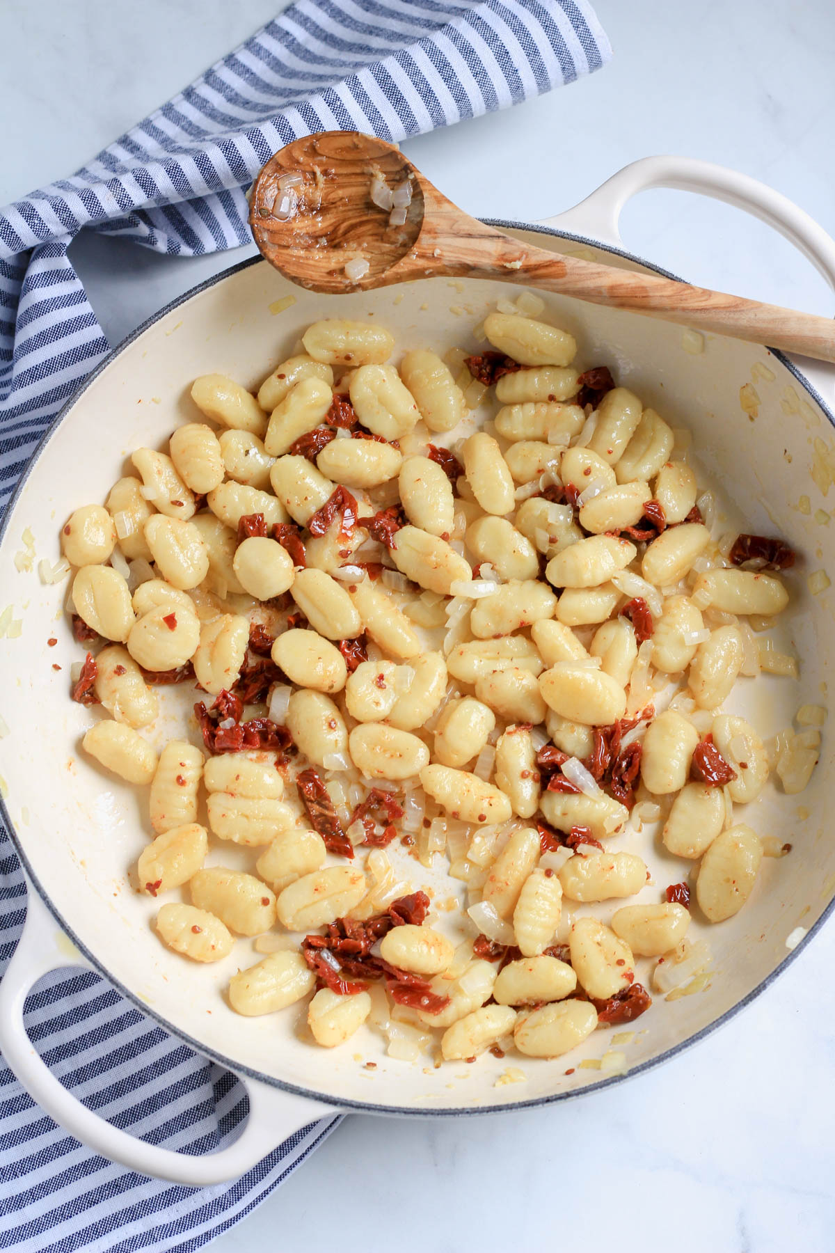 A white skillet with gnocchi, sun dried tomatoes, and a wooden spoon.