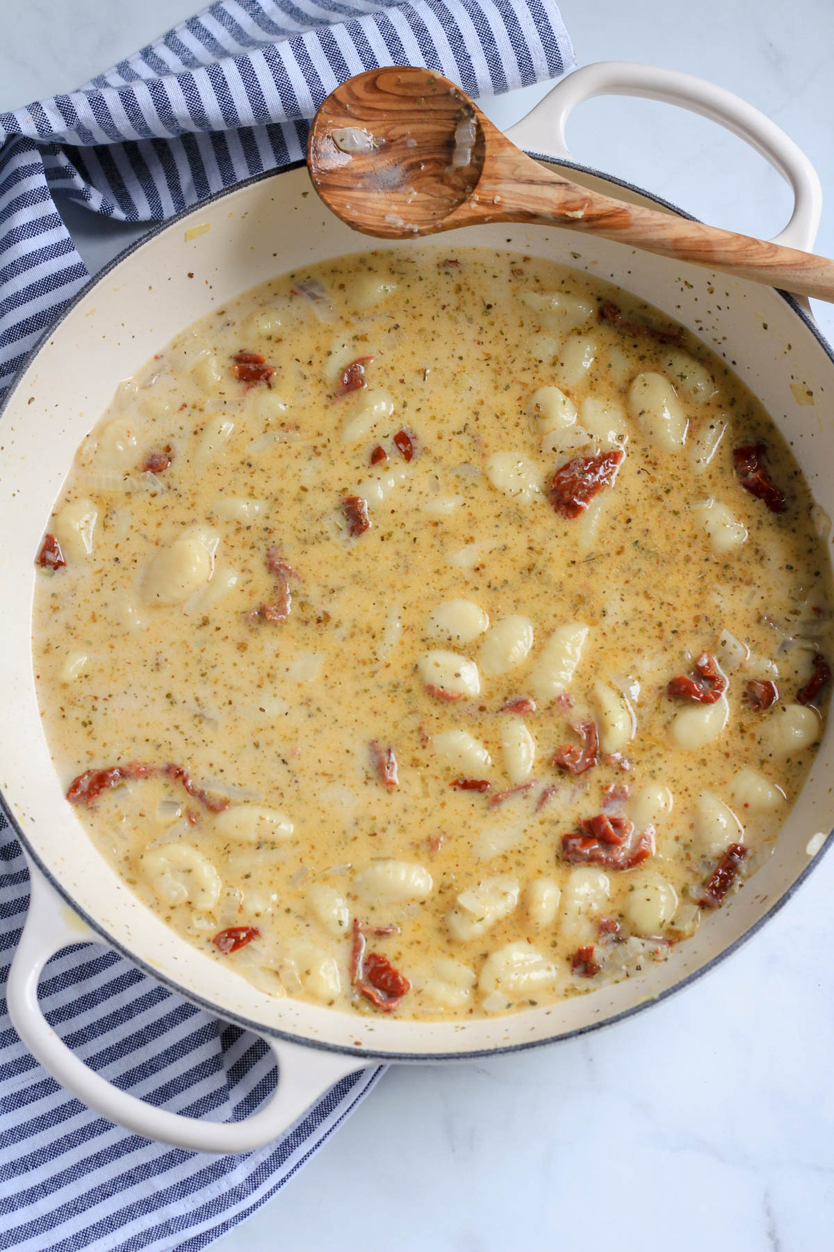 A white skillet with gnocchi and all of the liquids for the sauce with a wooden spoon.