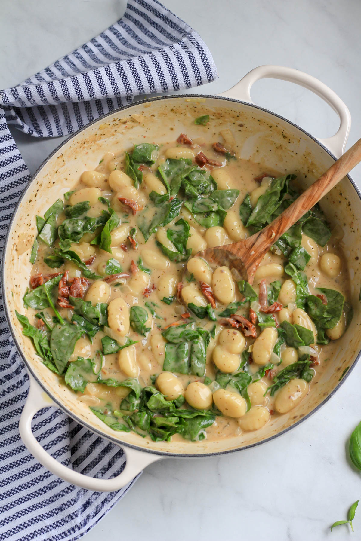 A white skillet on a white and blue towel with vegan gnocchi in a creamy sun dried tomato sauce with a wooden spoon.