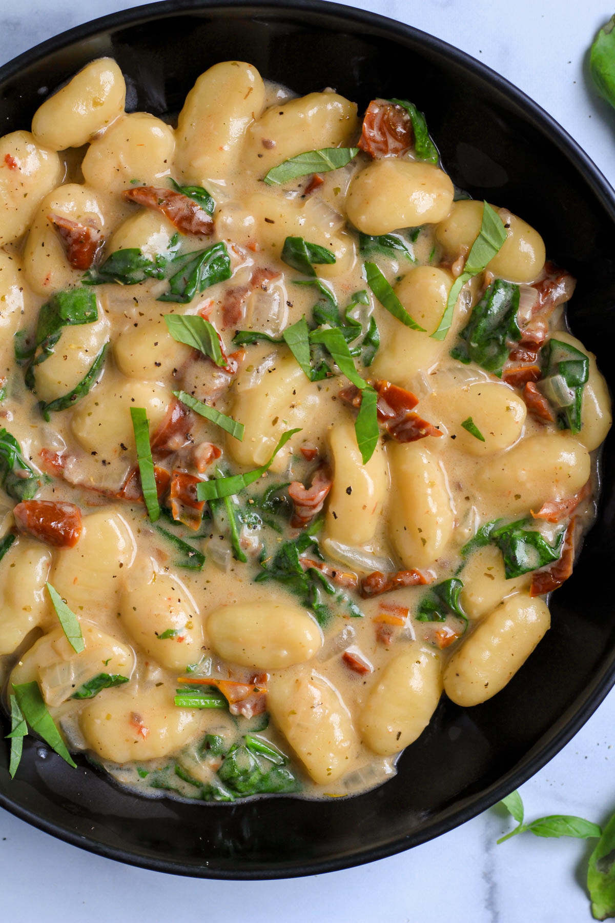 A close up of a black bowl of vegan gnocchi with sun dried tomato sauce on a white counter.