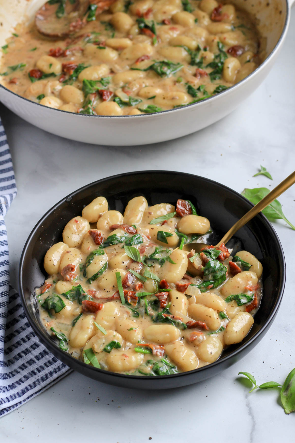 A black bowl of vegan gnocchi in front of a white skillet with creamy vegan gnocchi.