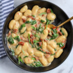 A black bowl of creamy vegan gnocchi topped with fresh basil and a gold spoon to the top right.