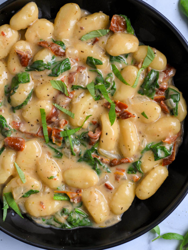 A close up of a black bowl of vegan gnocchi with sun dried tomato sauce on a white counter.