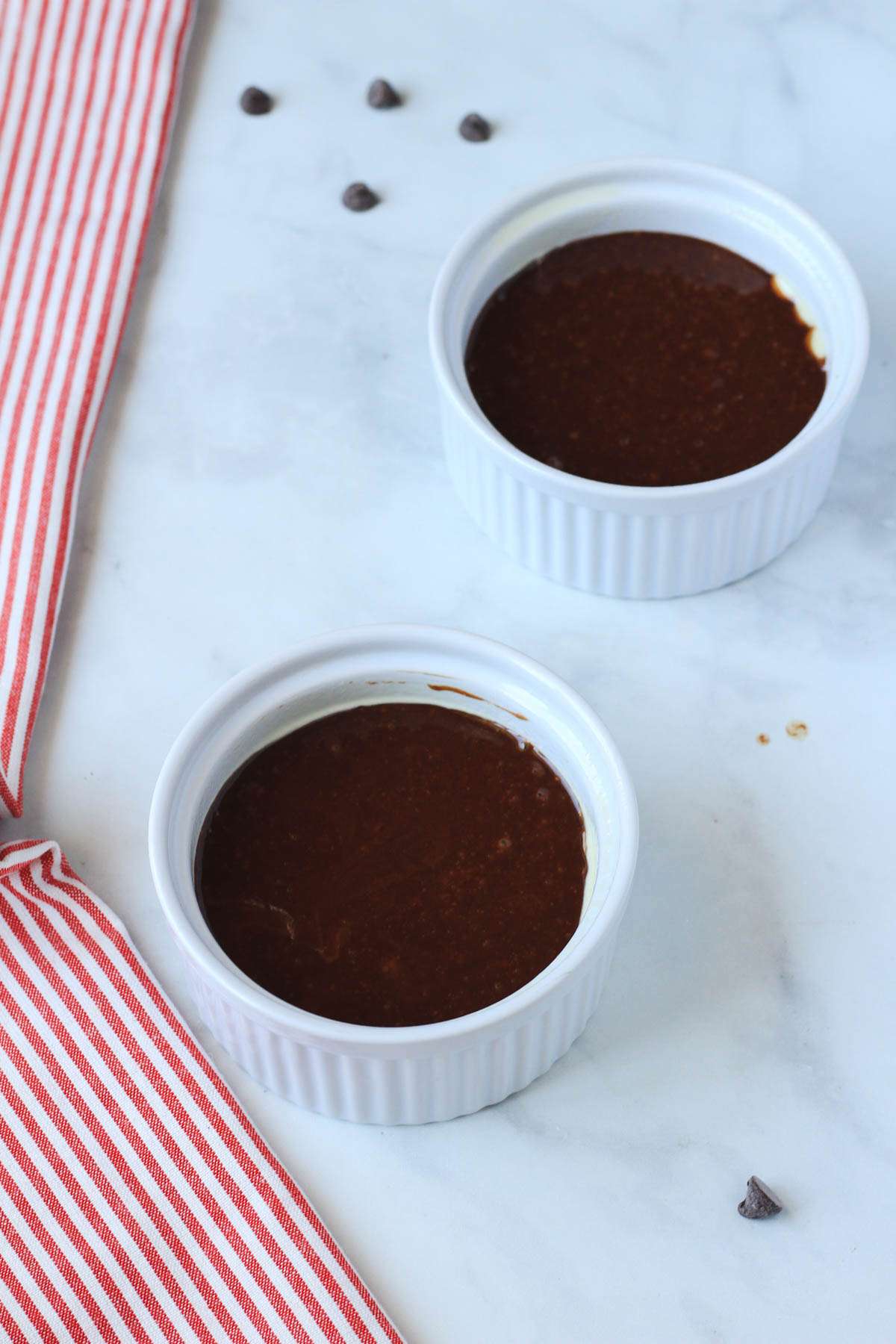 Two white ramekins filled with the dairy-free chocolate lava cakes on a white counter with a red and white striped dish towel to the left.