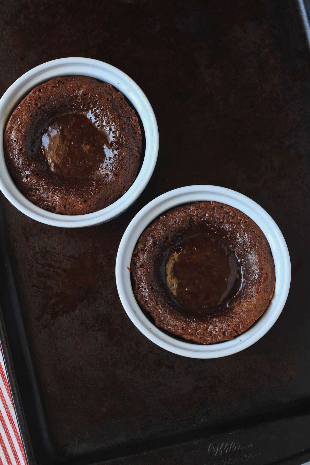 Two white ramekins with molten chocolate lava cakes after they bake on a baking sheet.