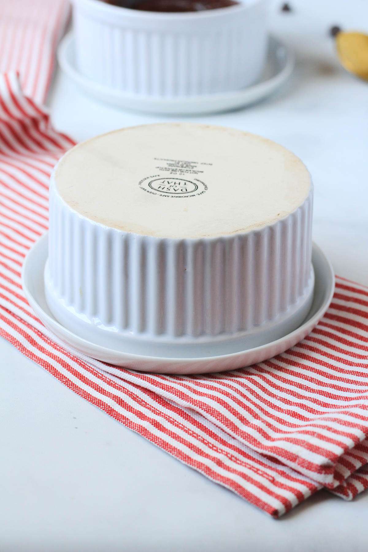 A white ramekin flipped on top of a white plate on a white and red striped towel.