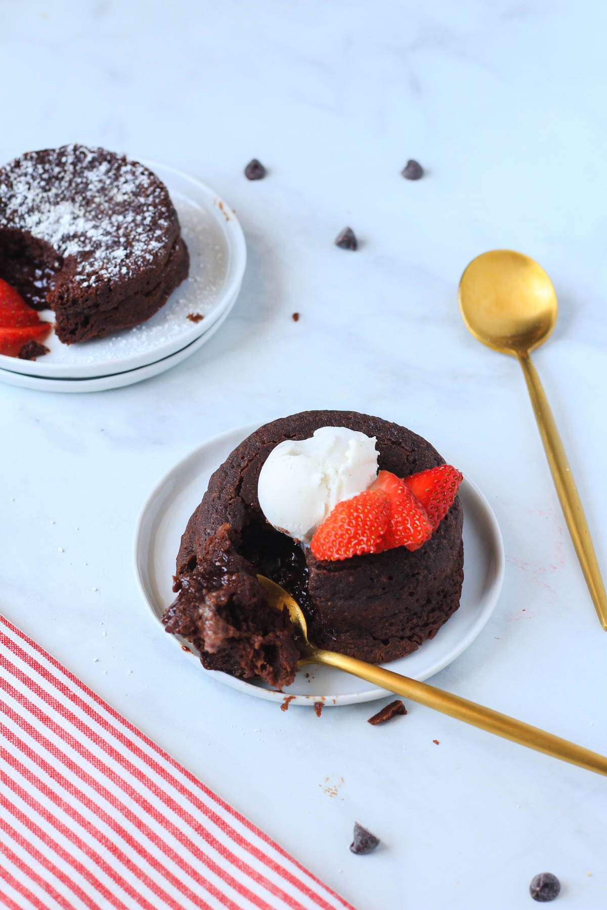 Two chocolate lava cakes on white plates the one in front topped with vegan ice cream with a bite on a golden spoon in the front.