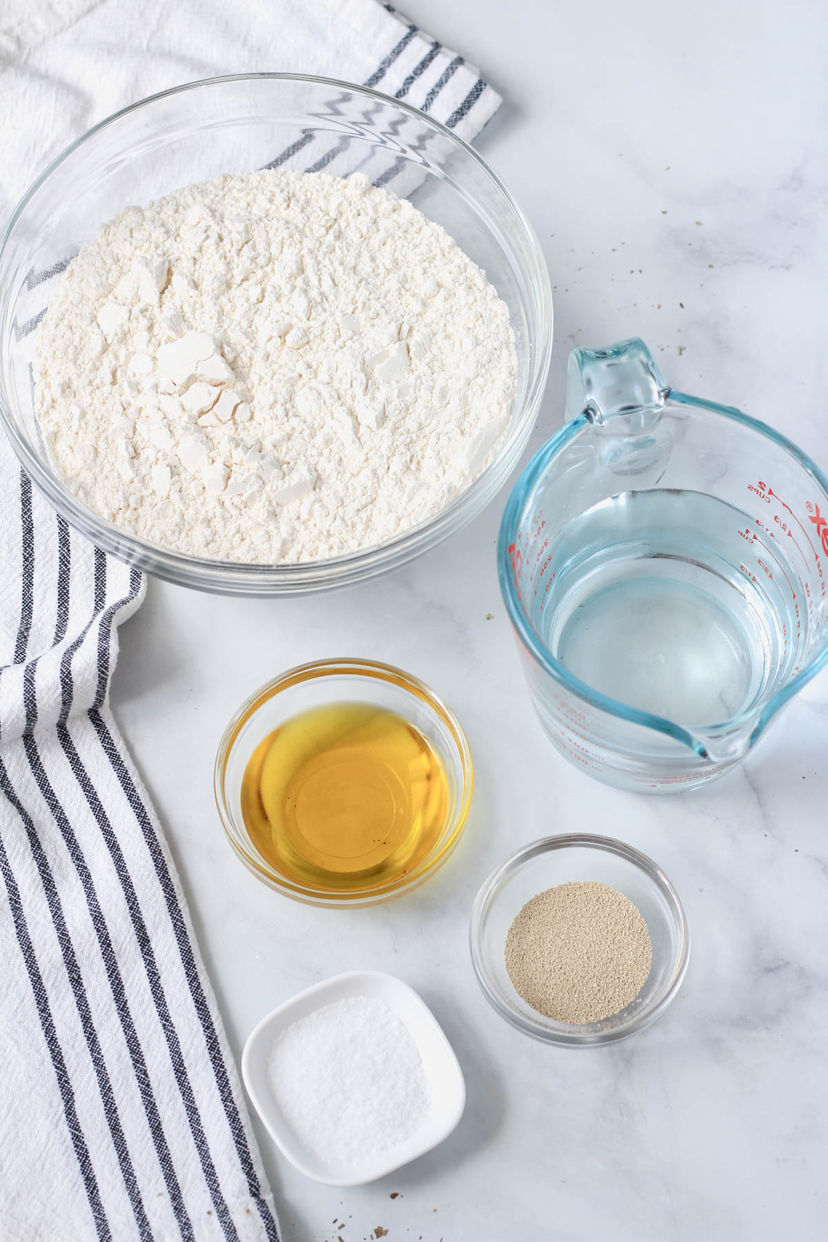 Ingredients for no-knead focaccia on a white counter with a blue and white striped towel to the right.