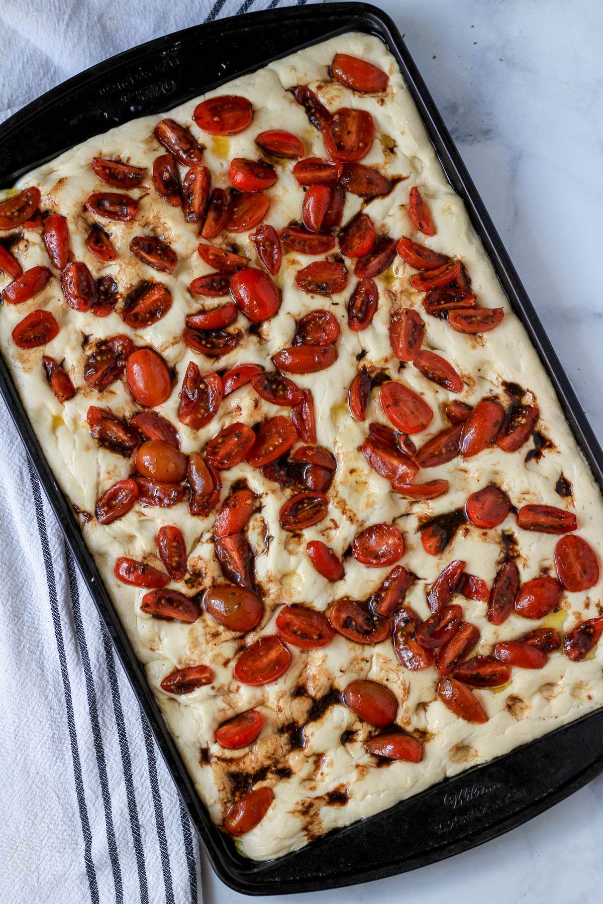 Focaccia with balsamic marinated tomatoes on top before baking in a rimmed baking sheet.