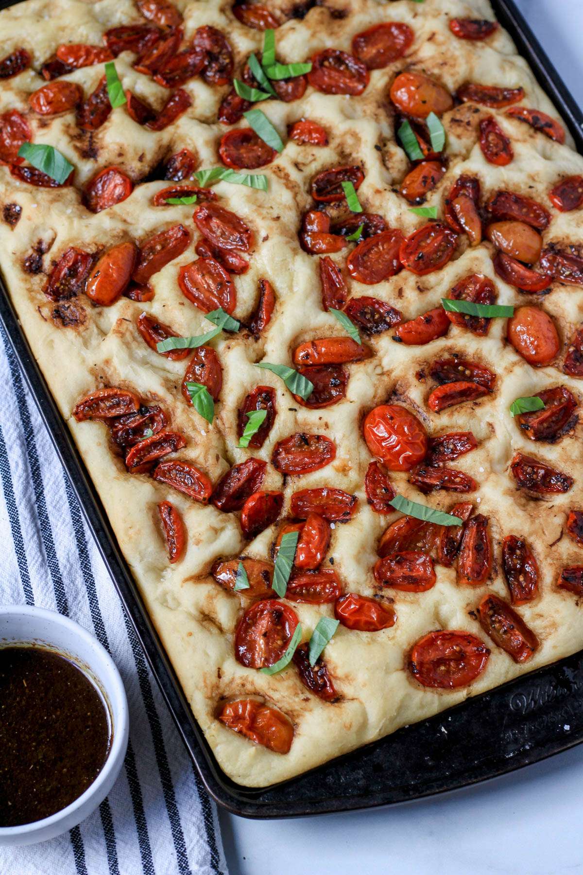 Marinated tomato focaccia topped with fresh basil on a rimmed baking sheet.