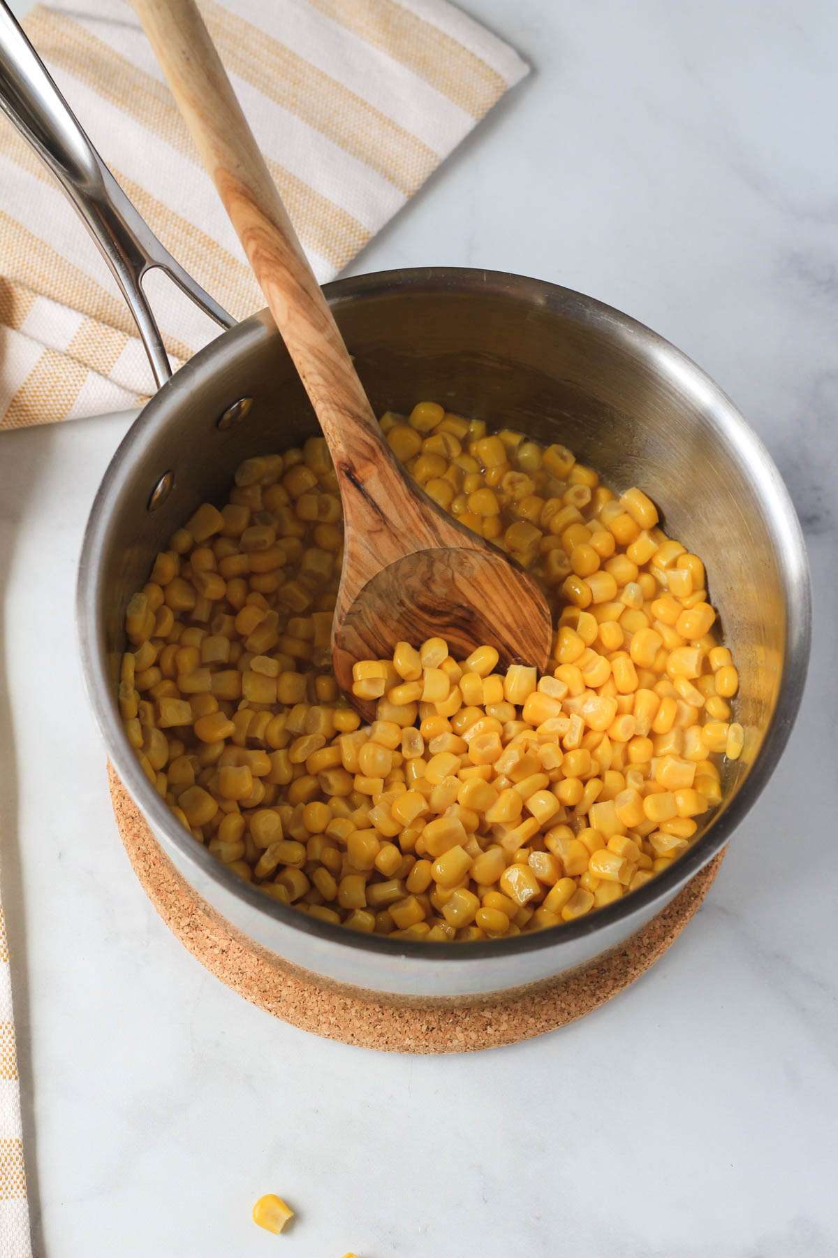 A silver sauce pan with a wooden spoon filled with corn.