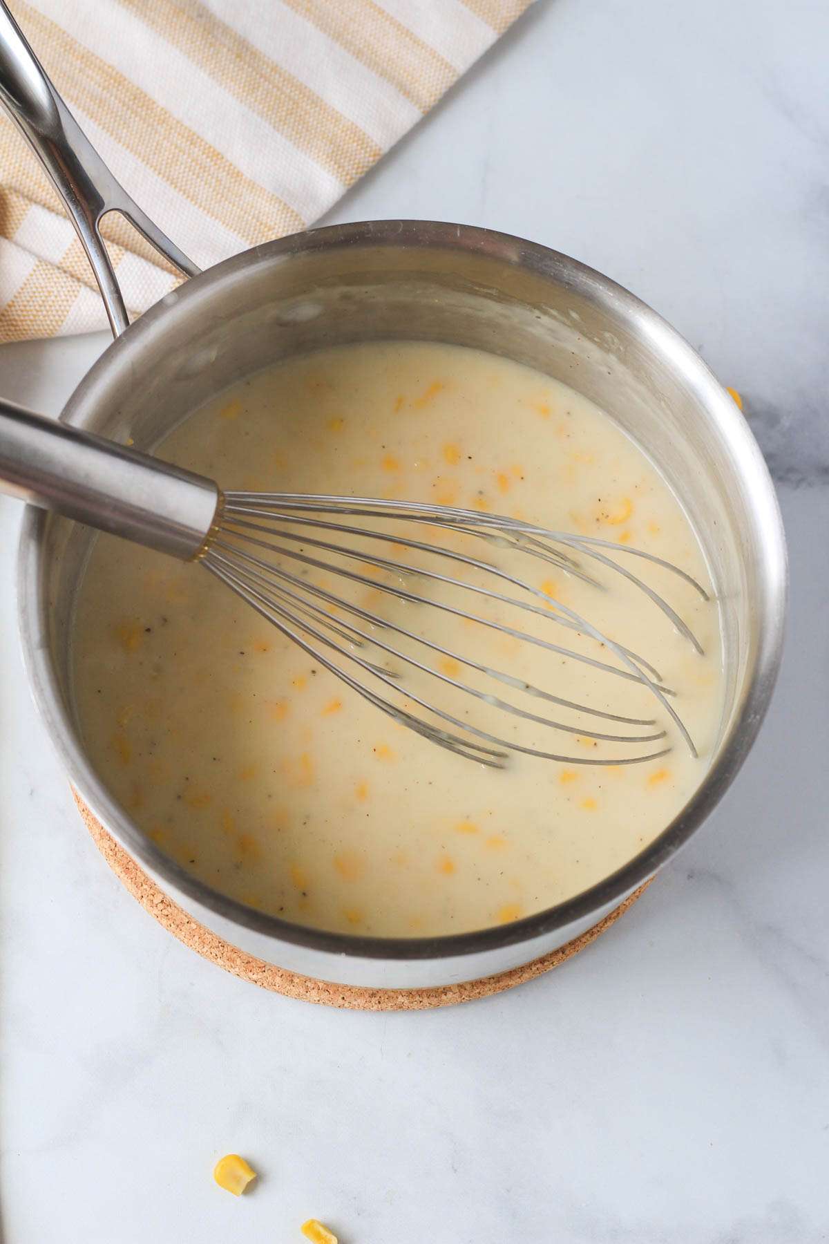 A wire whisk in a silver pan with creamed corn.