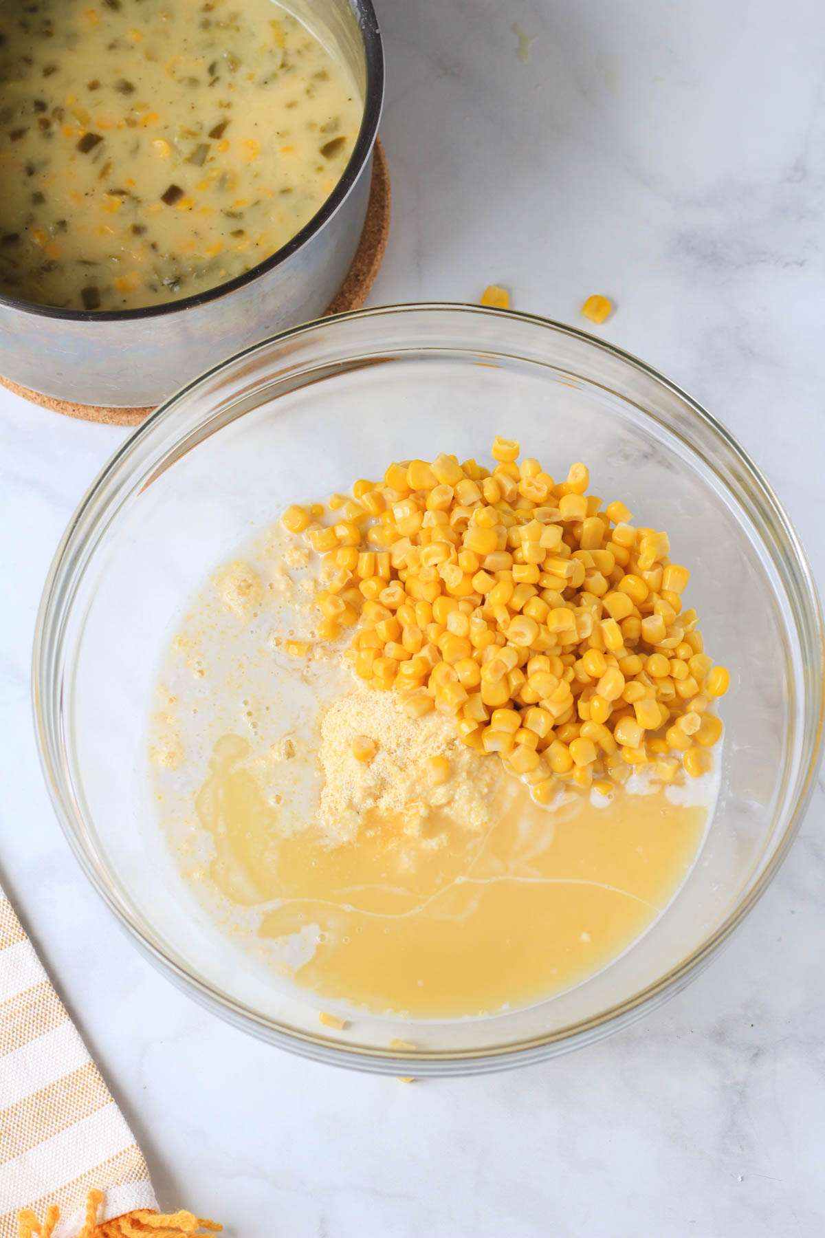 A glass mixing bowl with the ingredients for jalapeño corn casserole and a sauce pan with jalapeño creamed corn in the back.