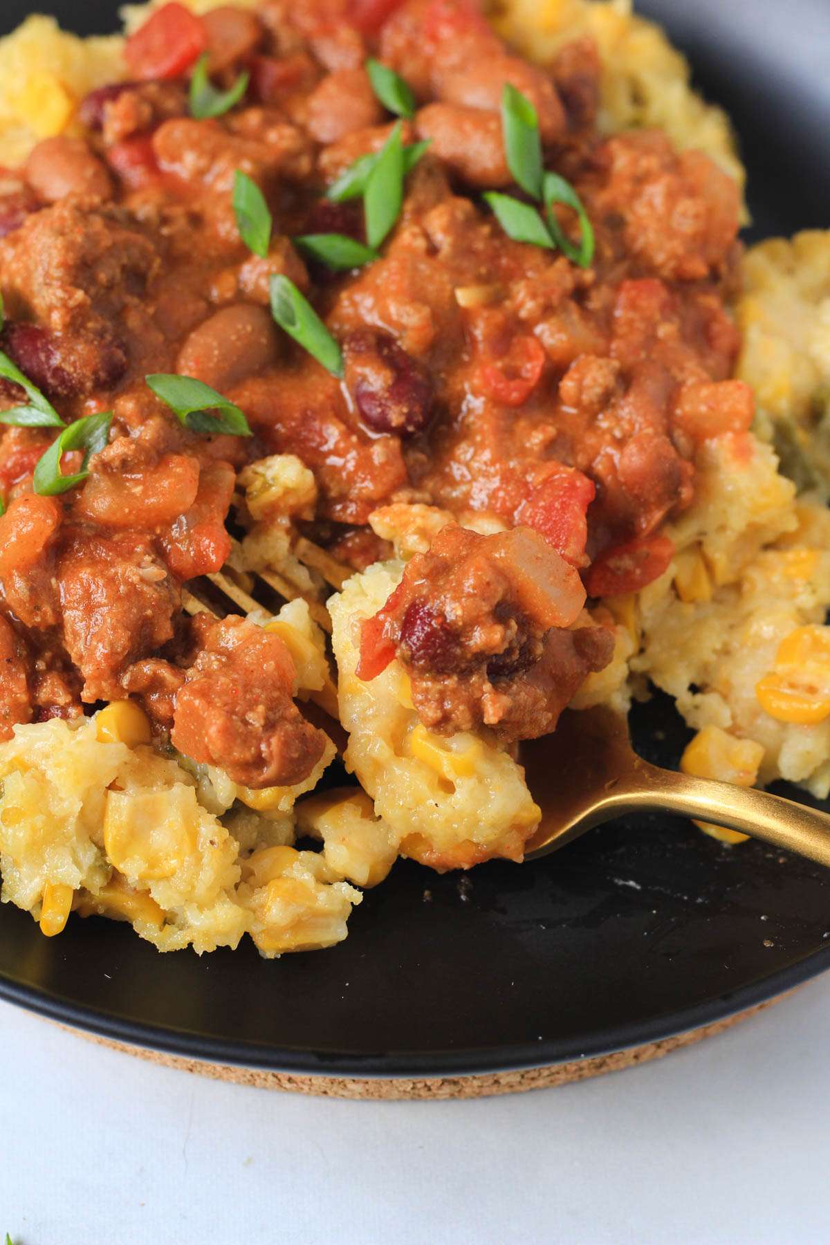 A gold fork topped with a bite of corn casserole and chili on a black plate.