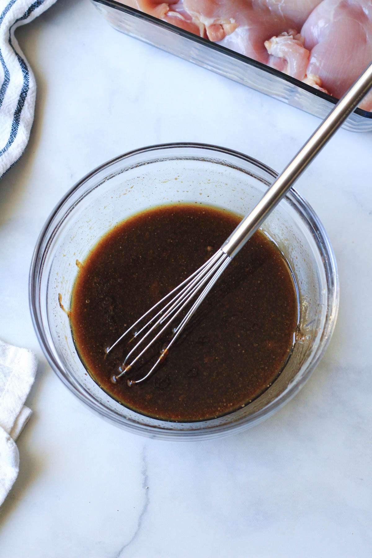 A glass bowl with a small silver whisk and the chicken thigh marinade all mixed together.
