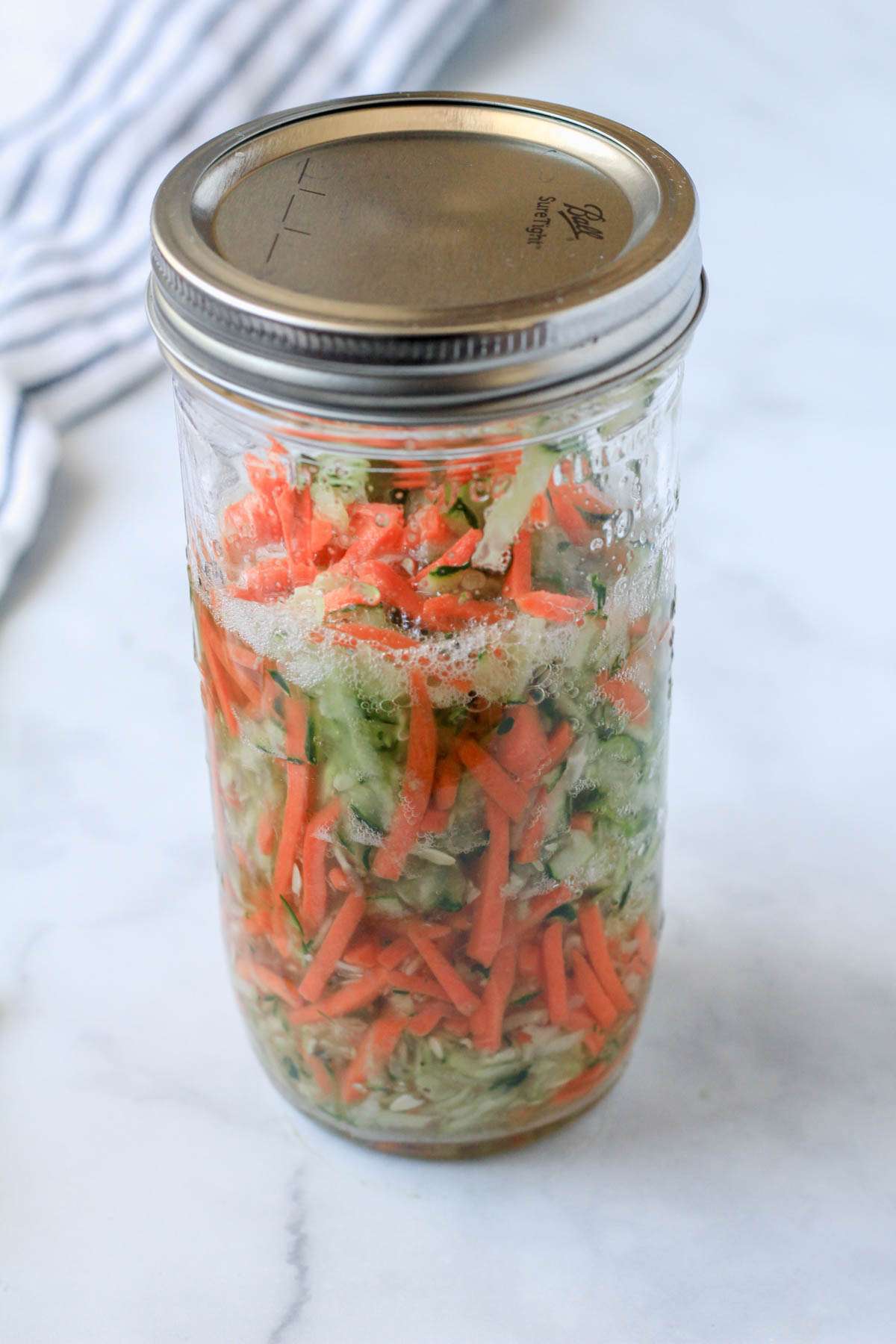 A tall mason jar with quick pickled cucumbers and carrots on a white counter.