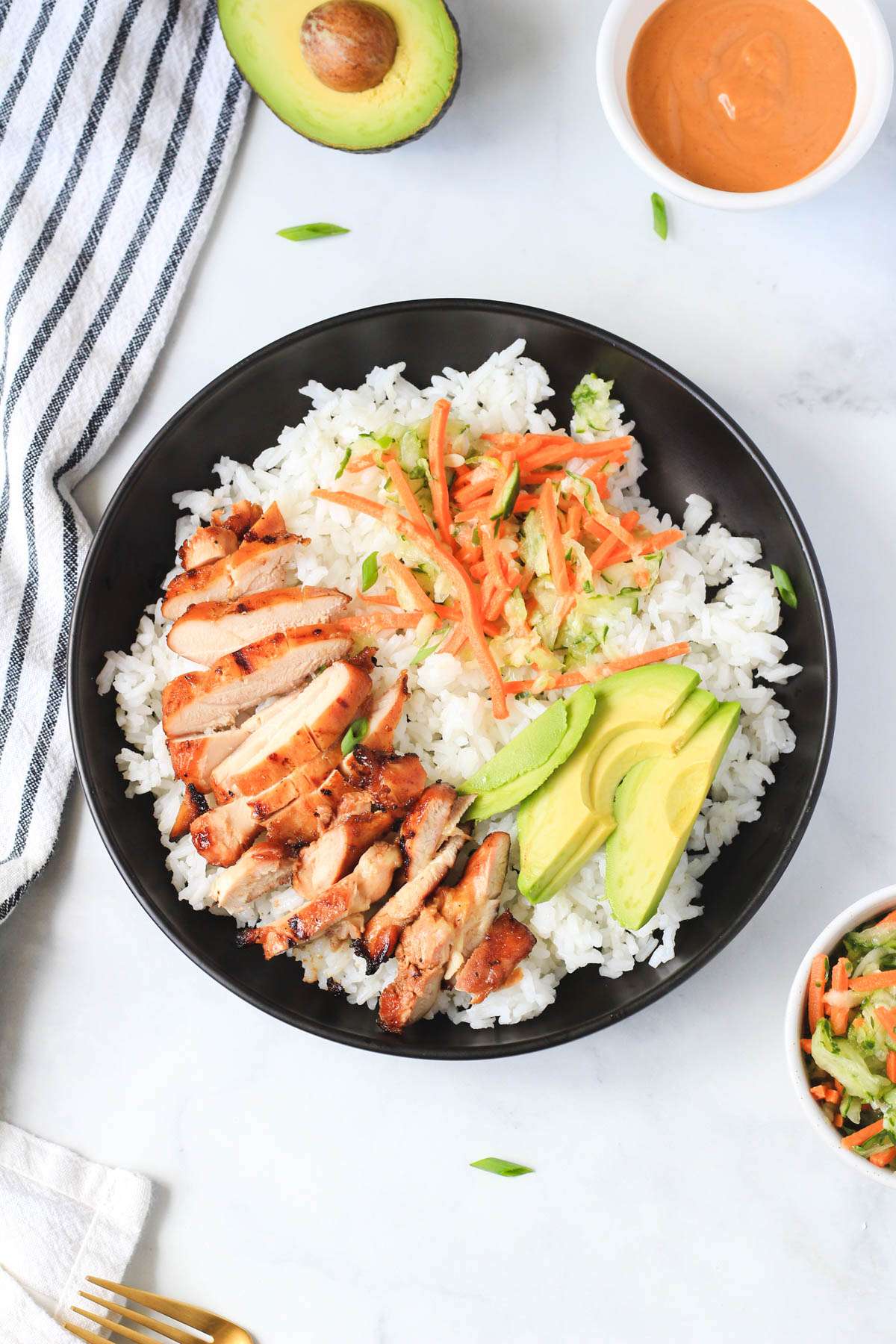A black bowl with soy-marinated chicken topping sticky rice with avocado and pickled cucumbers and carrots on top.