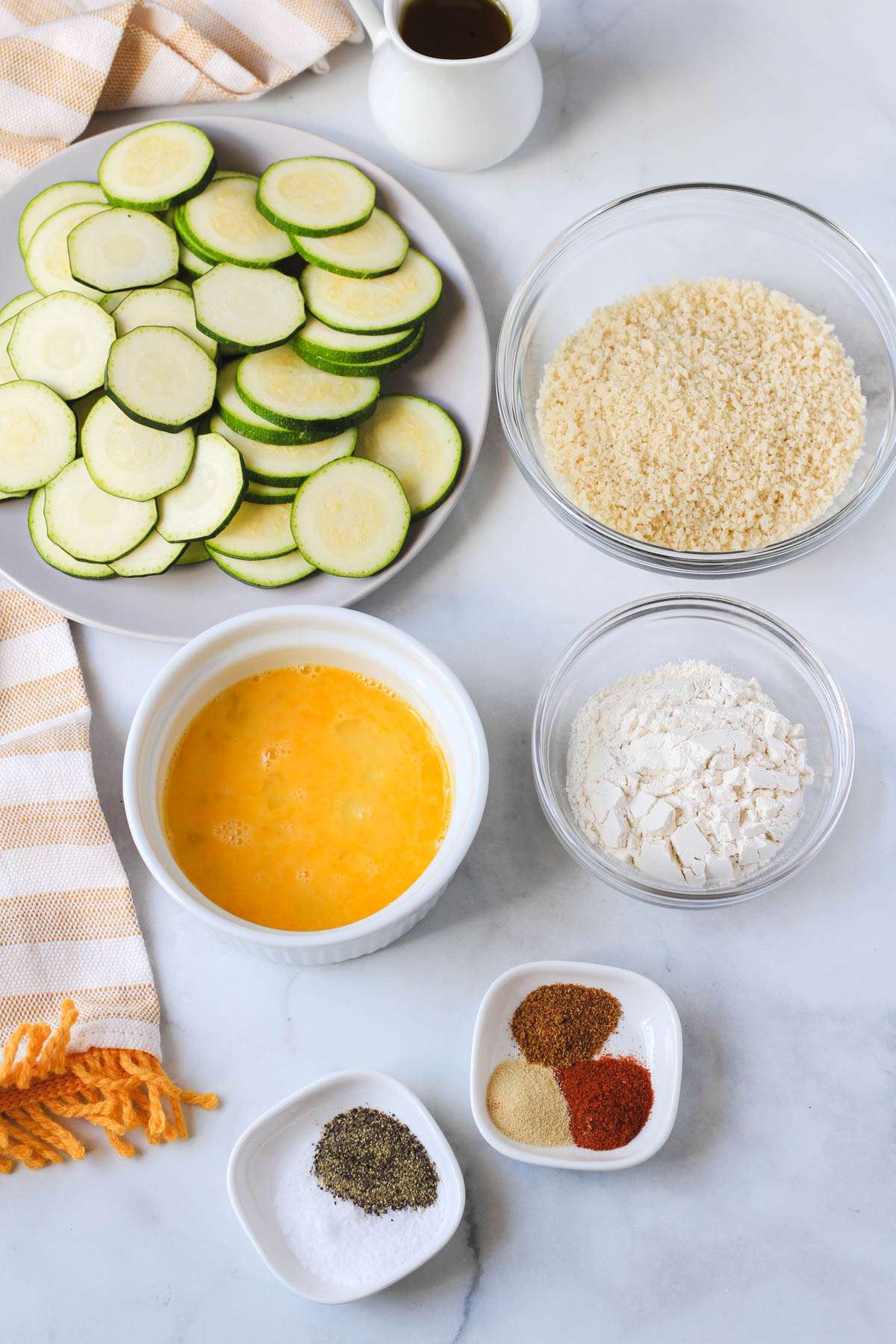 Ingredients for spicy air fryer zucchini bites on a white counter with a yellow and white striped dish towel.