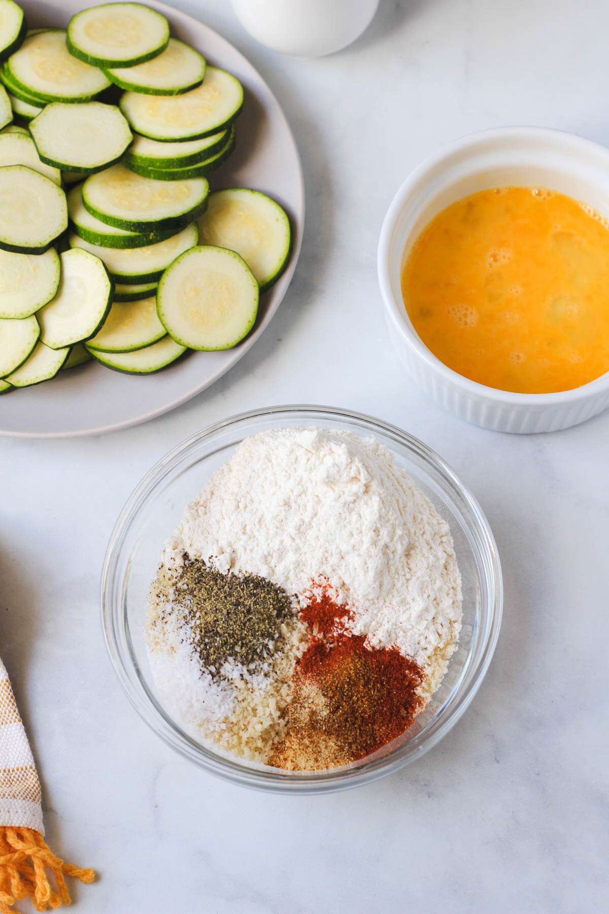 A glass bowl with all of the dry ingredients in the front and a bowl with a whisked egg in the back right and a plate of sliced zucchini in the back left.