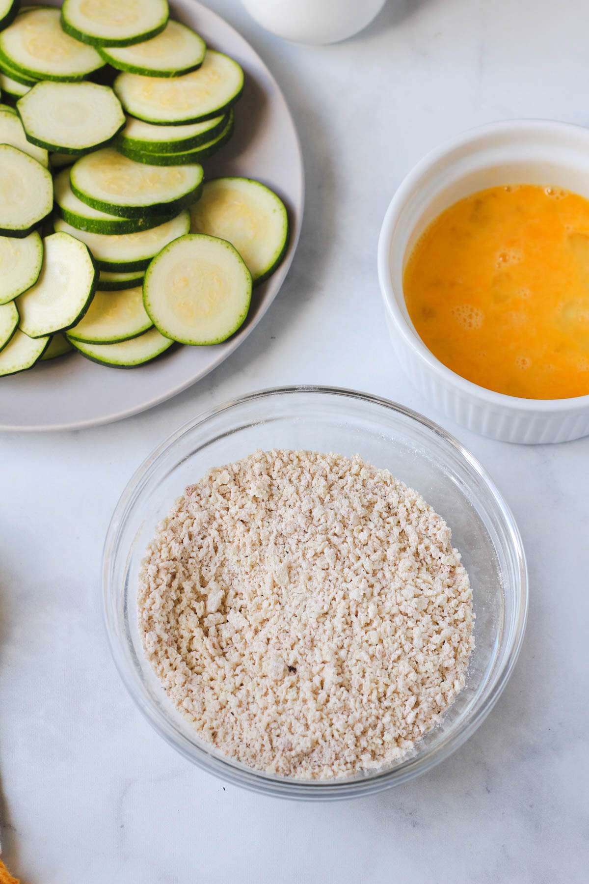 The bread crumb mixture in a glass bowl in the front and a plate of zucchini and a bowl with a whisked egg in the back.