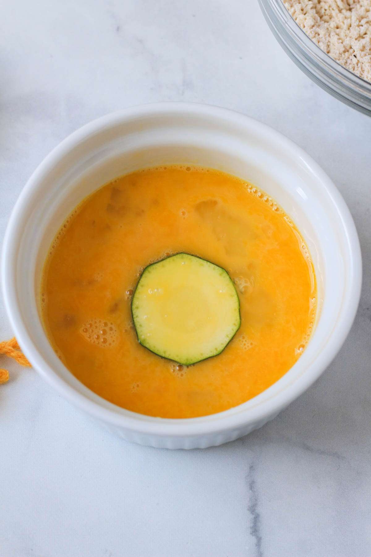 A white bowl with a whisked egg and a slice of zucchini in the center.