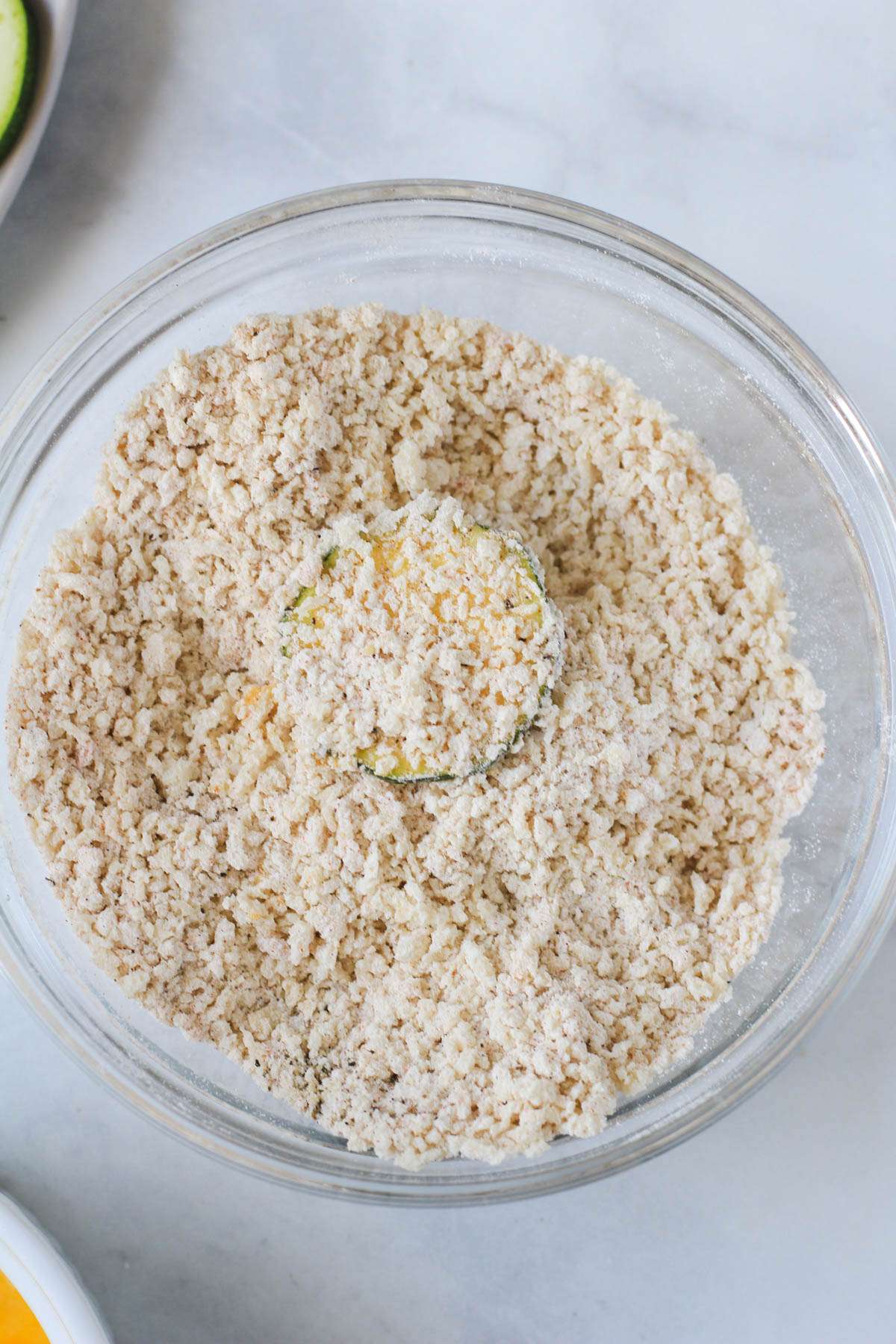 A glass bowl with the breading mixture and a slice of zucchini coated in breading in the center.