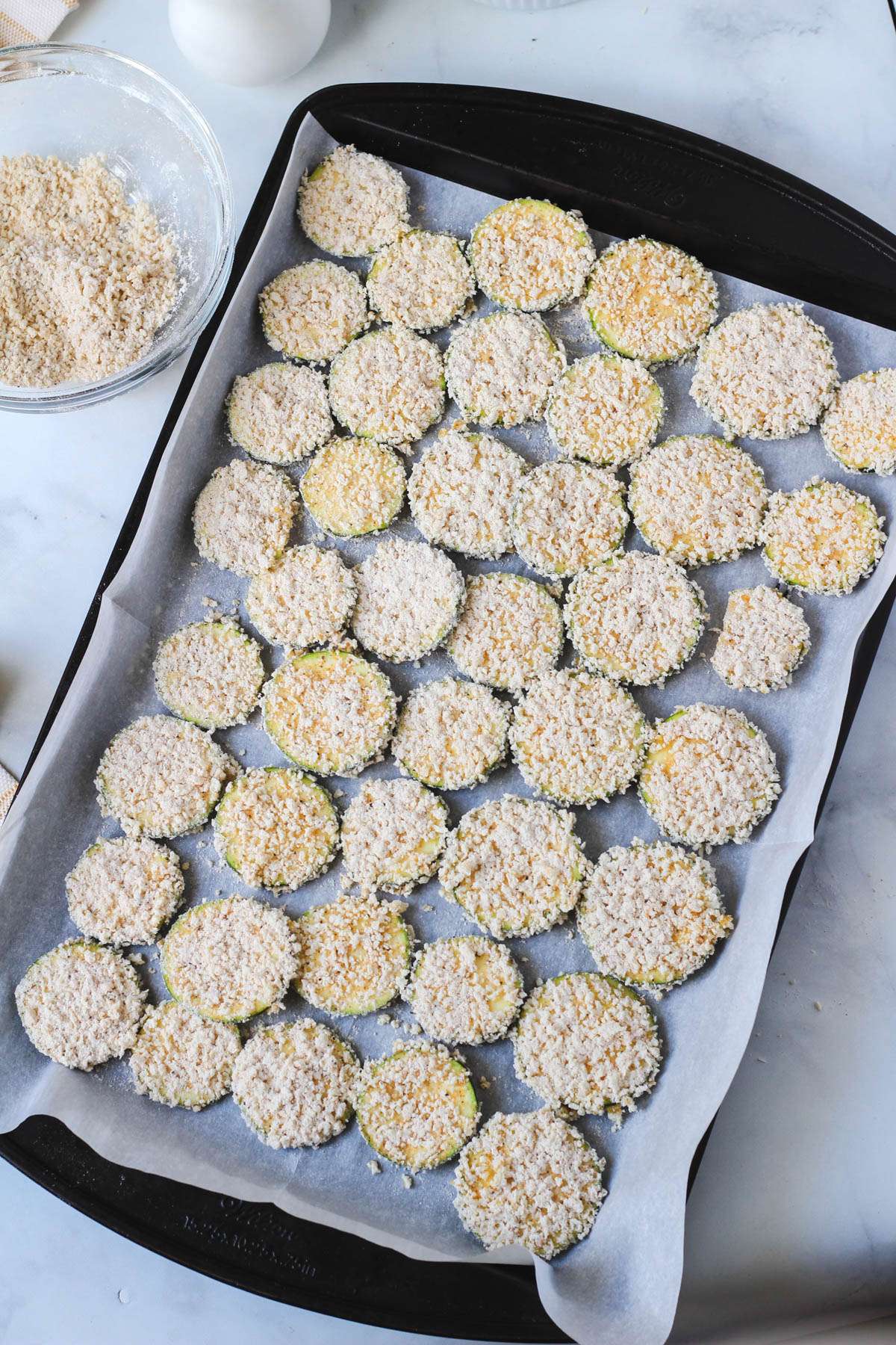 A parchment paper lined baking sheet covered in breaded zucchini bites before getting olive oil.