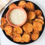 A top down photo of a black plate topped with crispy air fryer zucchini bites and a small bowl of dairy-free ranch dip in the top left corner of the plate.