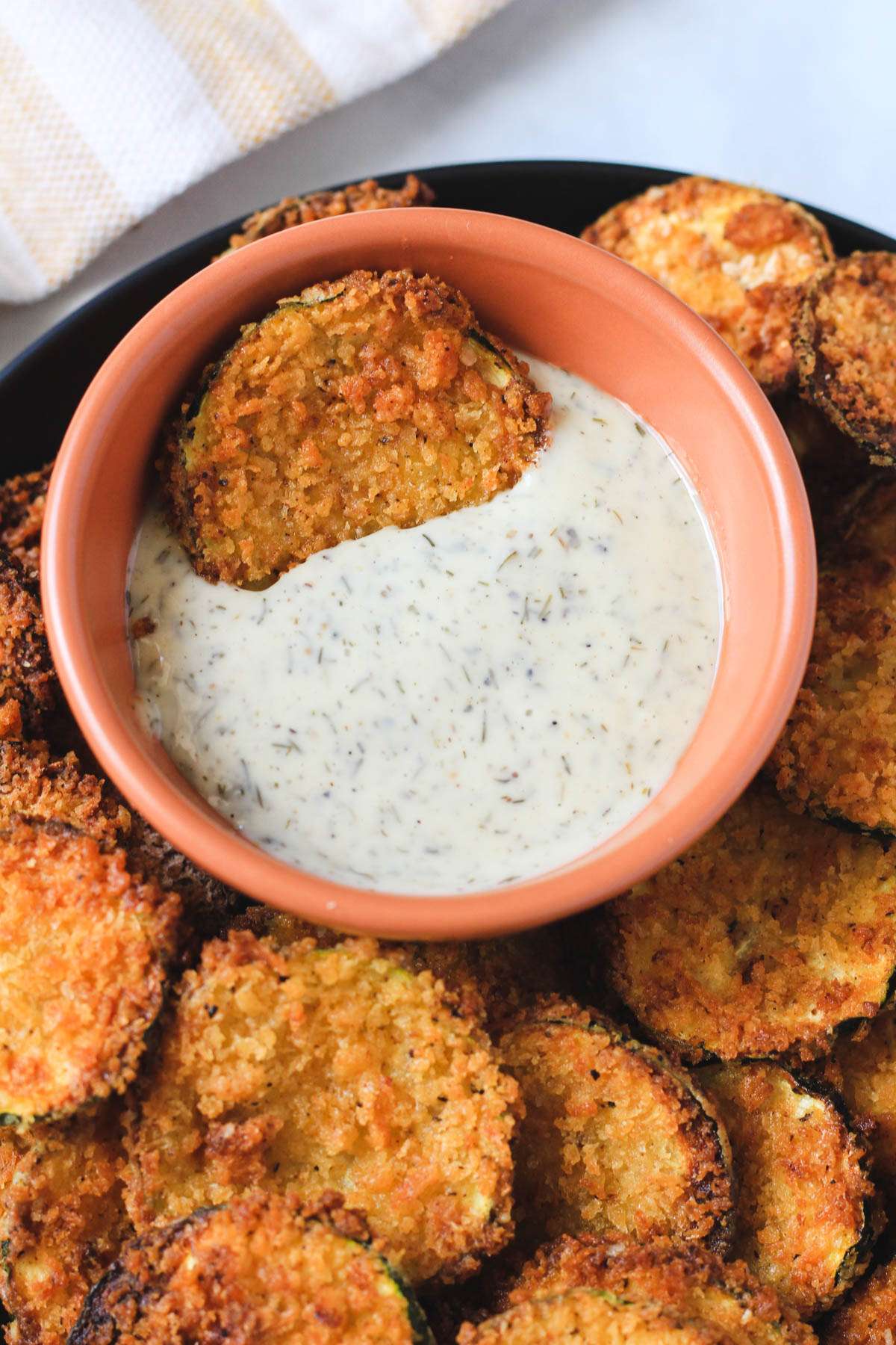 An orange bowl filled with dairy-free ranch dressing with a spicy air fryer zucchini bite in the bowl and more spicy zucchini bites around the bowl.