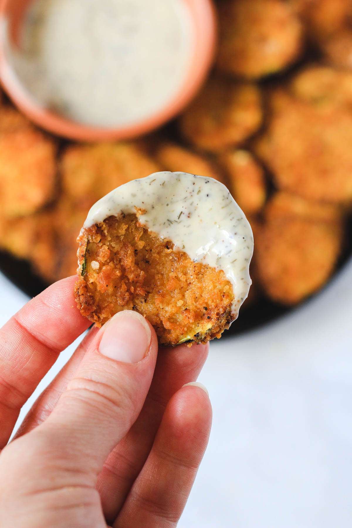 A hand holding a spicy air fryer zucchini bite dipped in dairy-free ranch dressing.