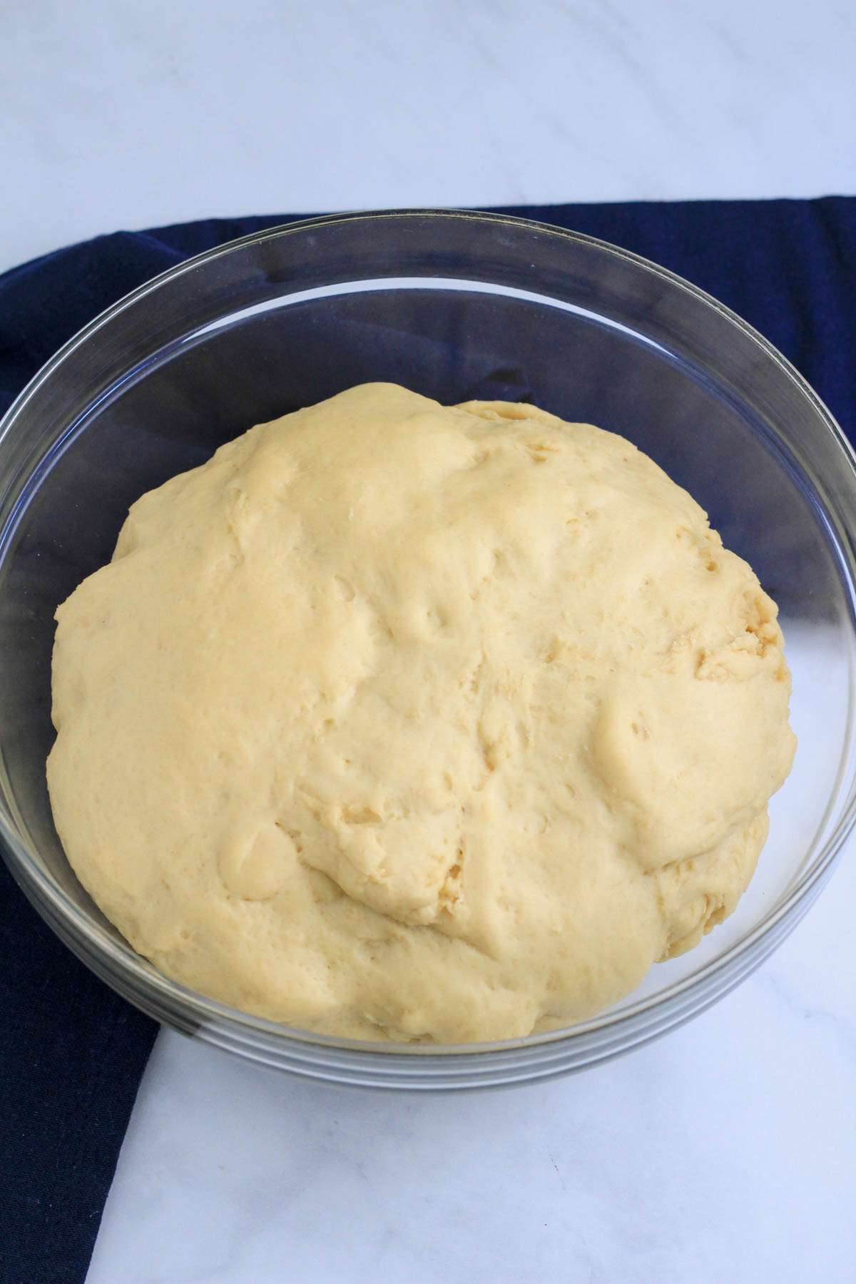 A glass bowl on a white counter with a blue towel with cinnamon roll dough after doubling in size.