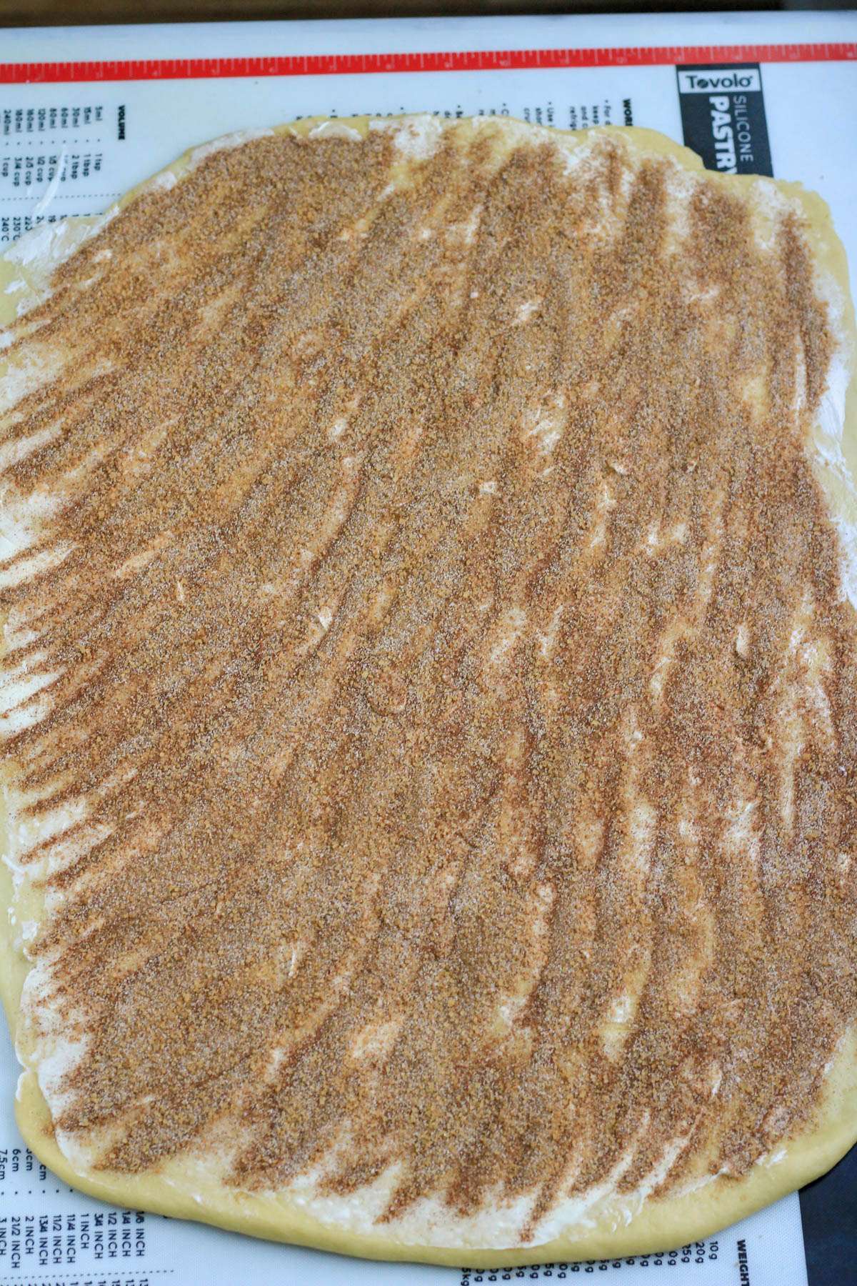 A pastry mat with cinnamon roll dough rolled out and topped with butter and cinnamon sugar.