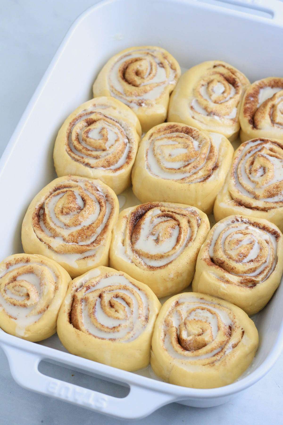 A white baking pan with cinnamon rolls after rising topped with coconut milk before baking.