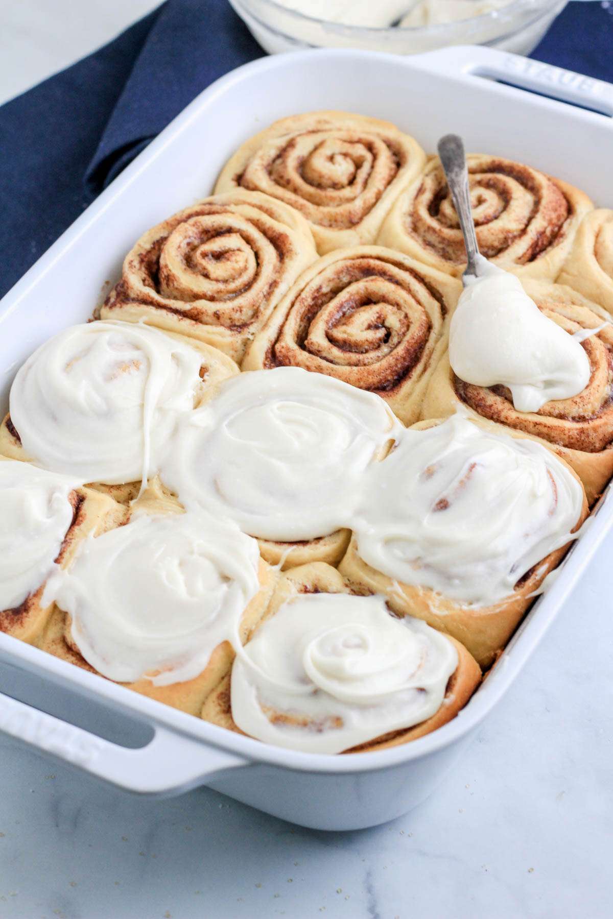 A white baking dish with dairy-free traditional cinnamon rolls while they are being frosted.