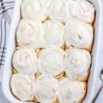 A close up of frosted dairy-free cinnamon rolls in a white baking dish on a white counter.