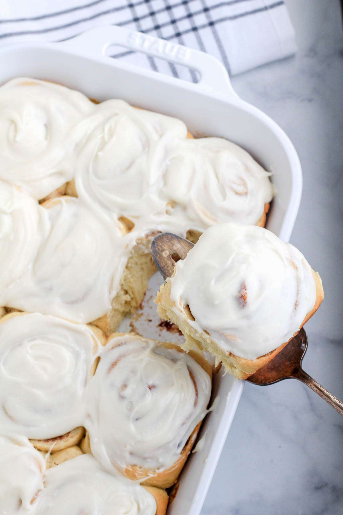 A pie server with a cinnamon roll on top above the white baking dish filled with cinnamon rolls.