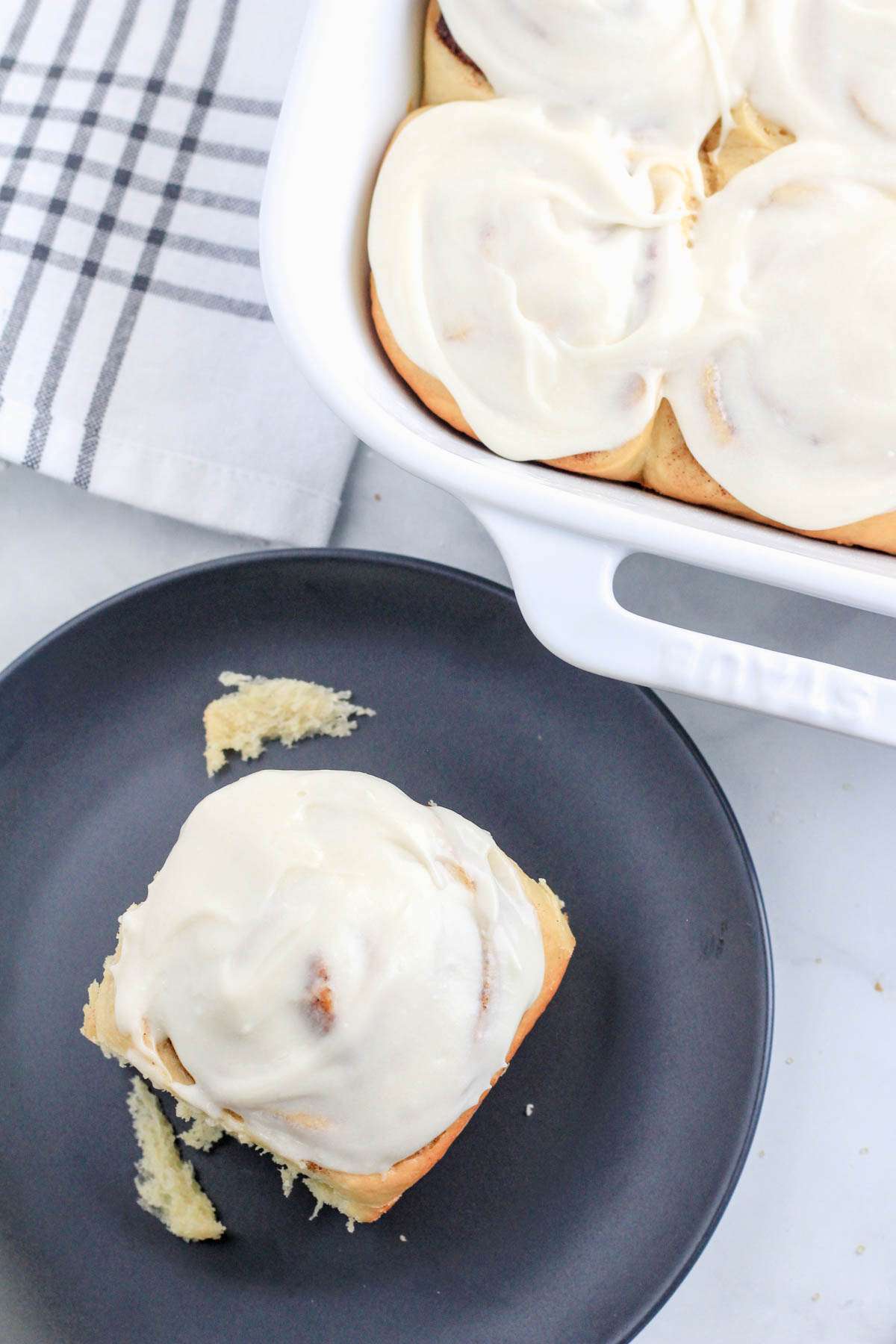 A blue plate with a traditional cinnamon roll on top and a white baking dish with cinnamon rolls in the top right corner.