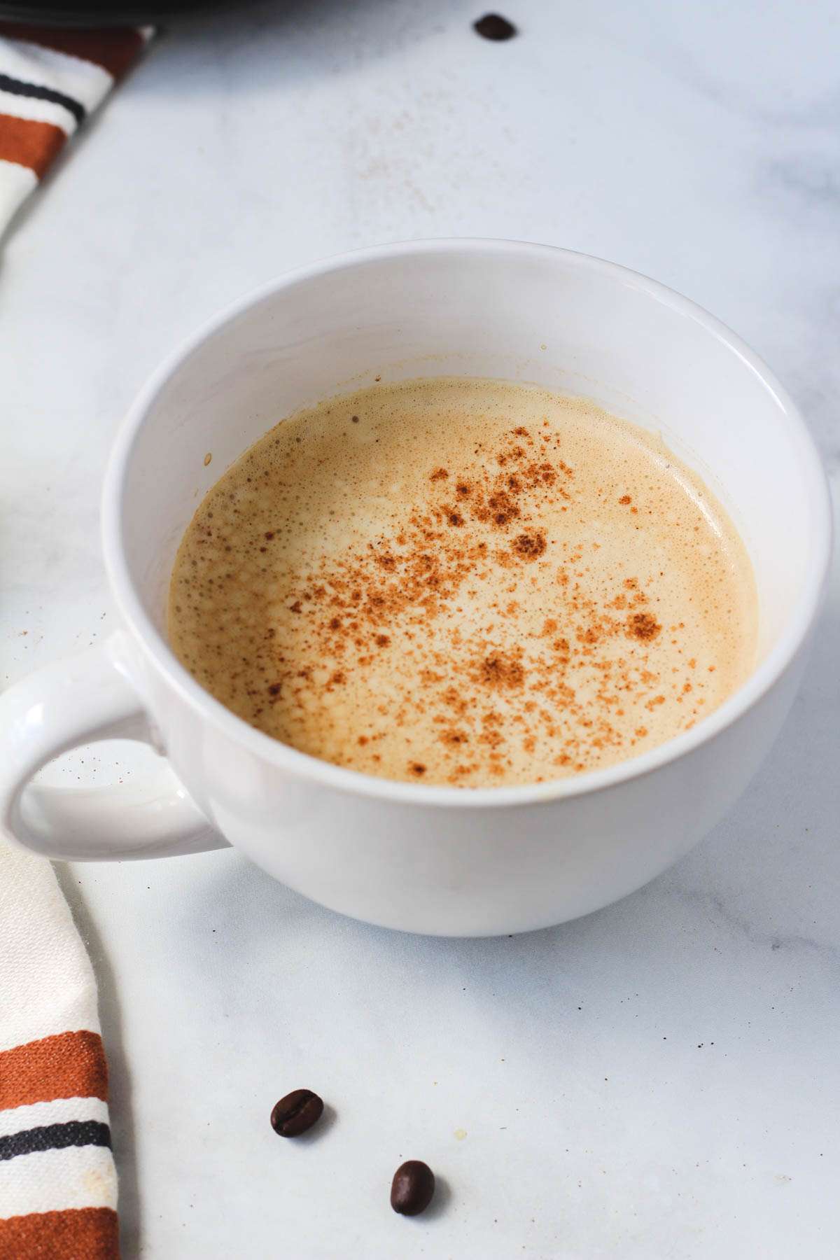 A white mug of vanilla protein coffee on a white counter with some coffee beans on the counter.