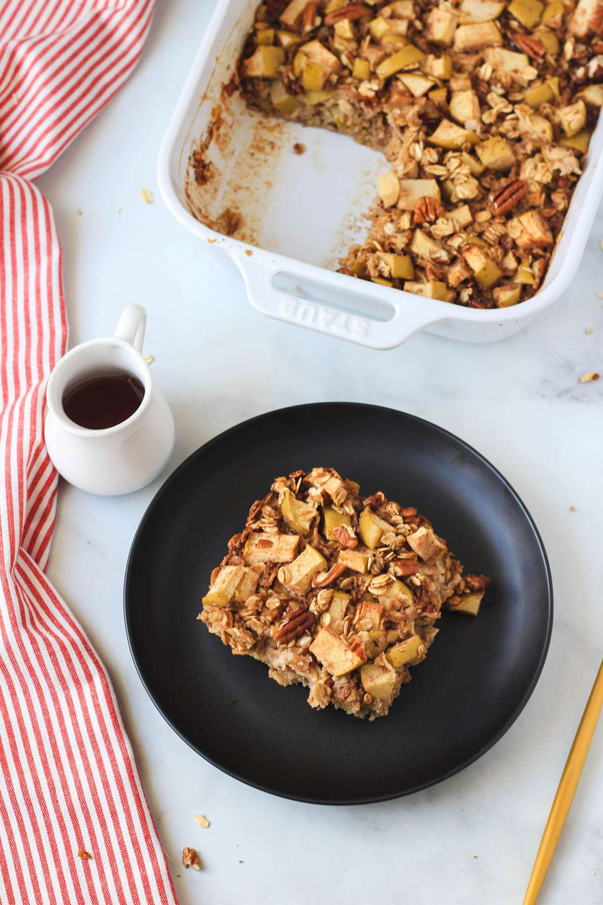 A plate with a slice of apple pie baked oatmeal with a gold fork to the right and a white jar of maple syrup to the left.