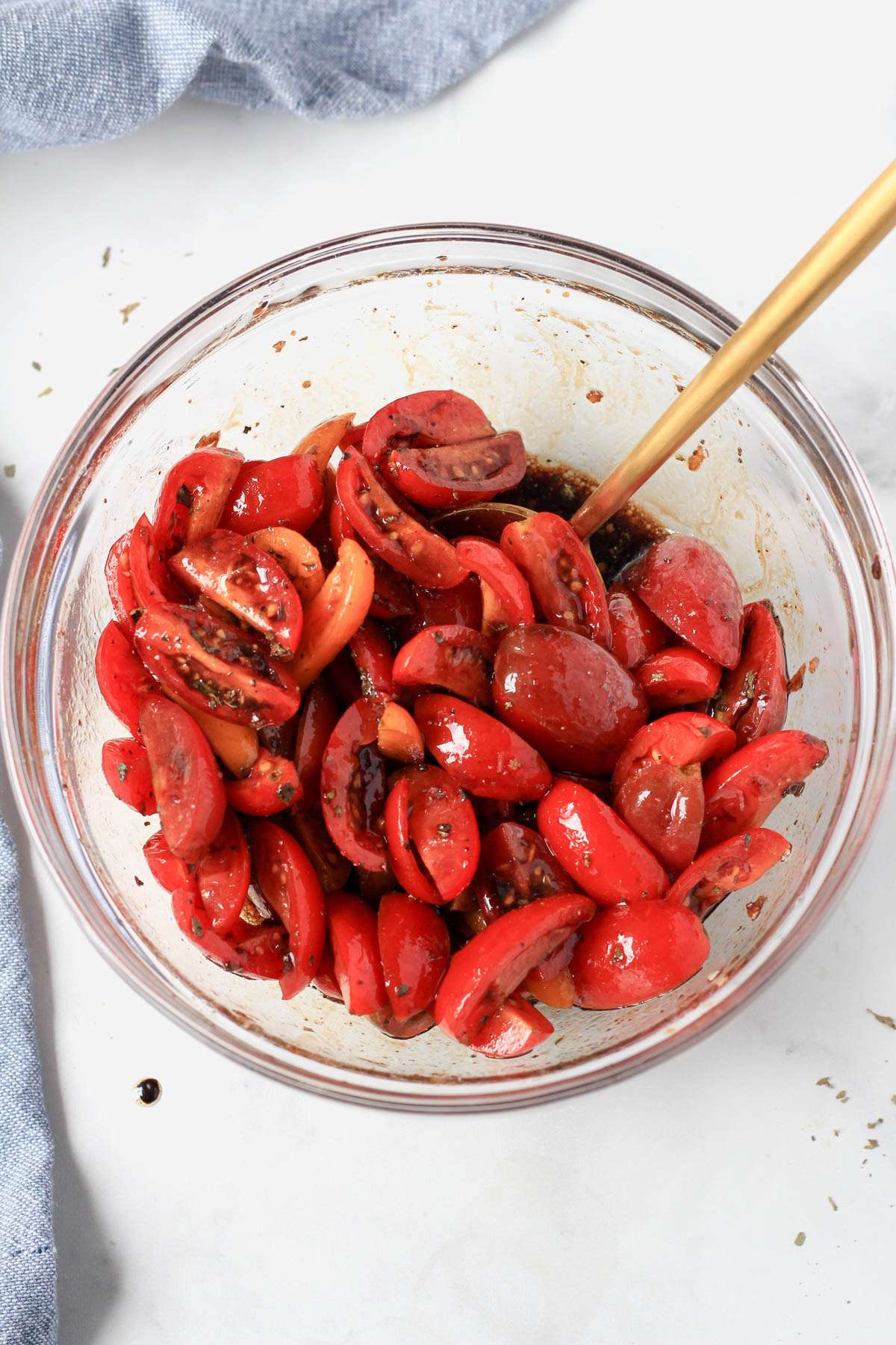 A glass bowl of balsamic marinated tomatoes with a gold spoon on the right side.